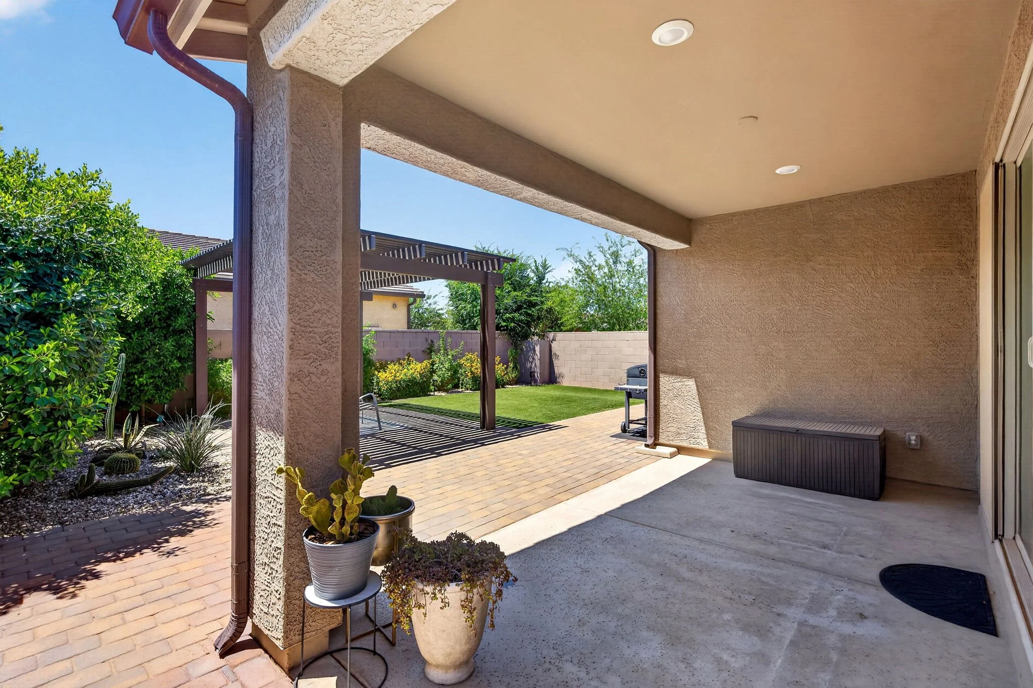 View from a covered patio to a backyard with a brick pathway, green grass, trees, and a fence, under a clear blue sky.