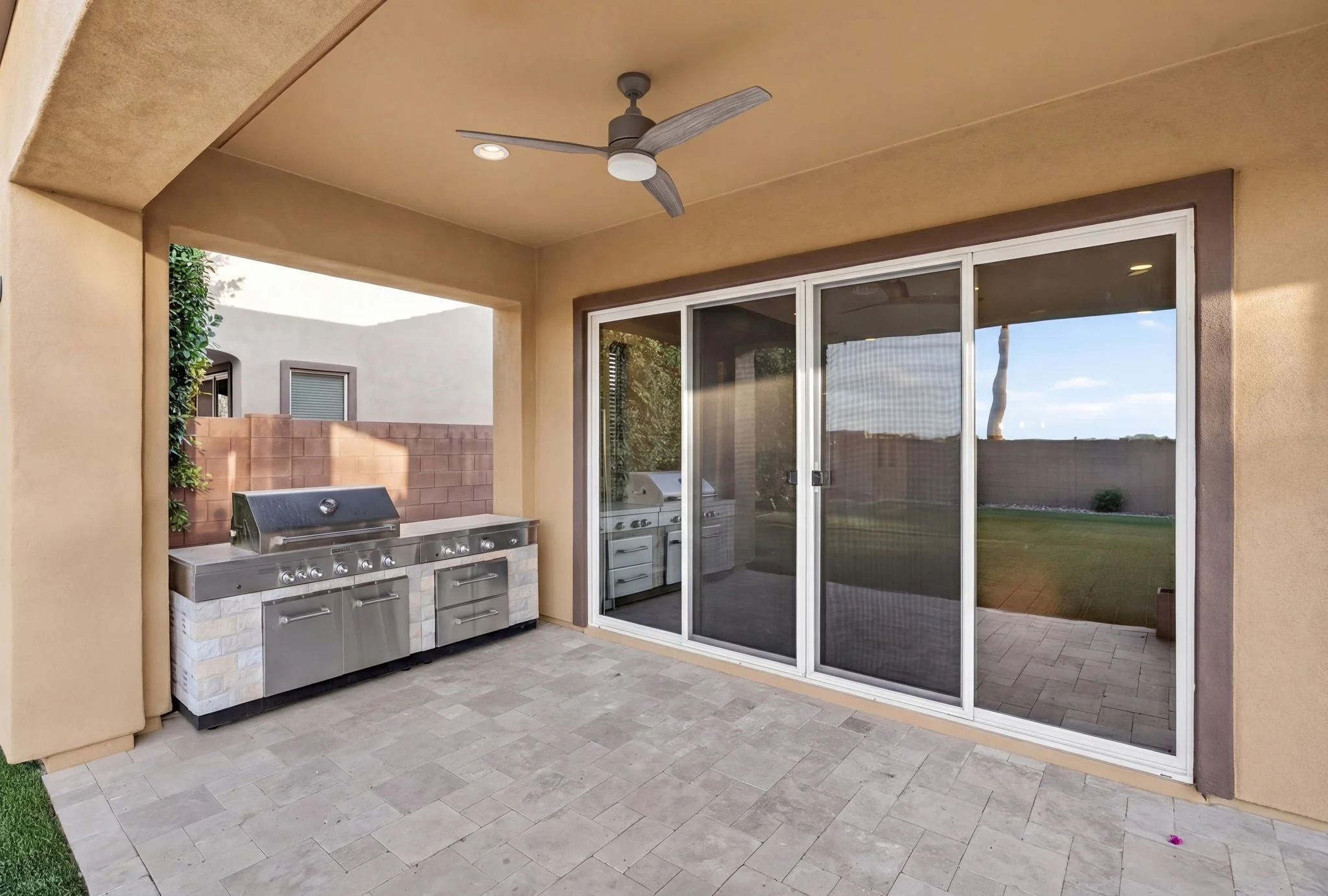 An outdoor patio area with a ceiling fan, a built-in grill, sliding glass doors leading inside, and a brick wall in the background.