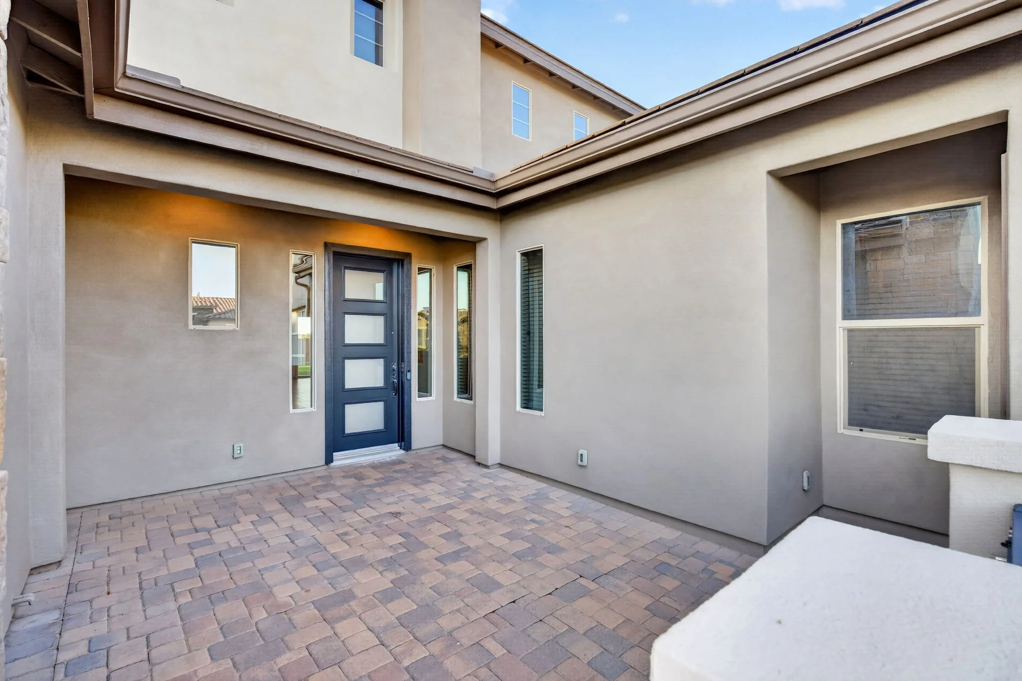 Backyard patio with brick pavers, a gray front door with glass panels, and beige stucco walls. There are multiple windows and a corner of a stone wall is visible on the left.