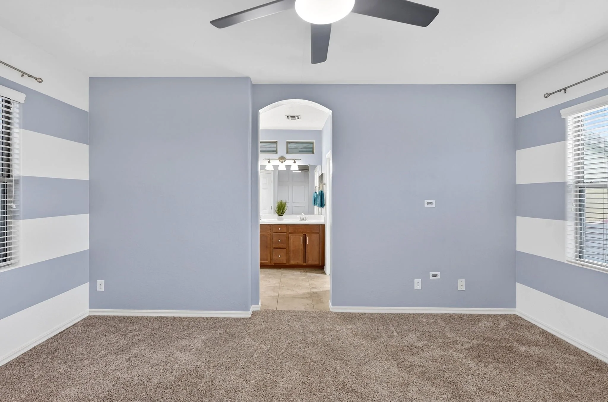 Empty living room with light blue walls, beige carpet, and two windows with blinds. An open doorway leads to a bathroom with a wooden vanity, a mirror, and a plant on the countertop.