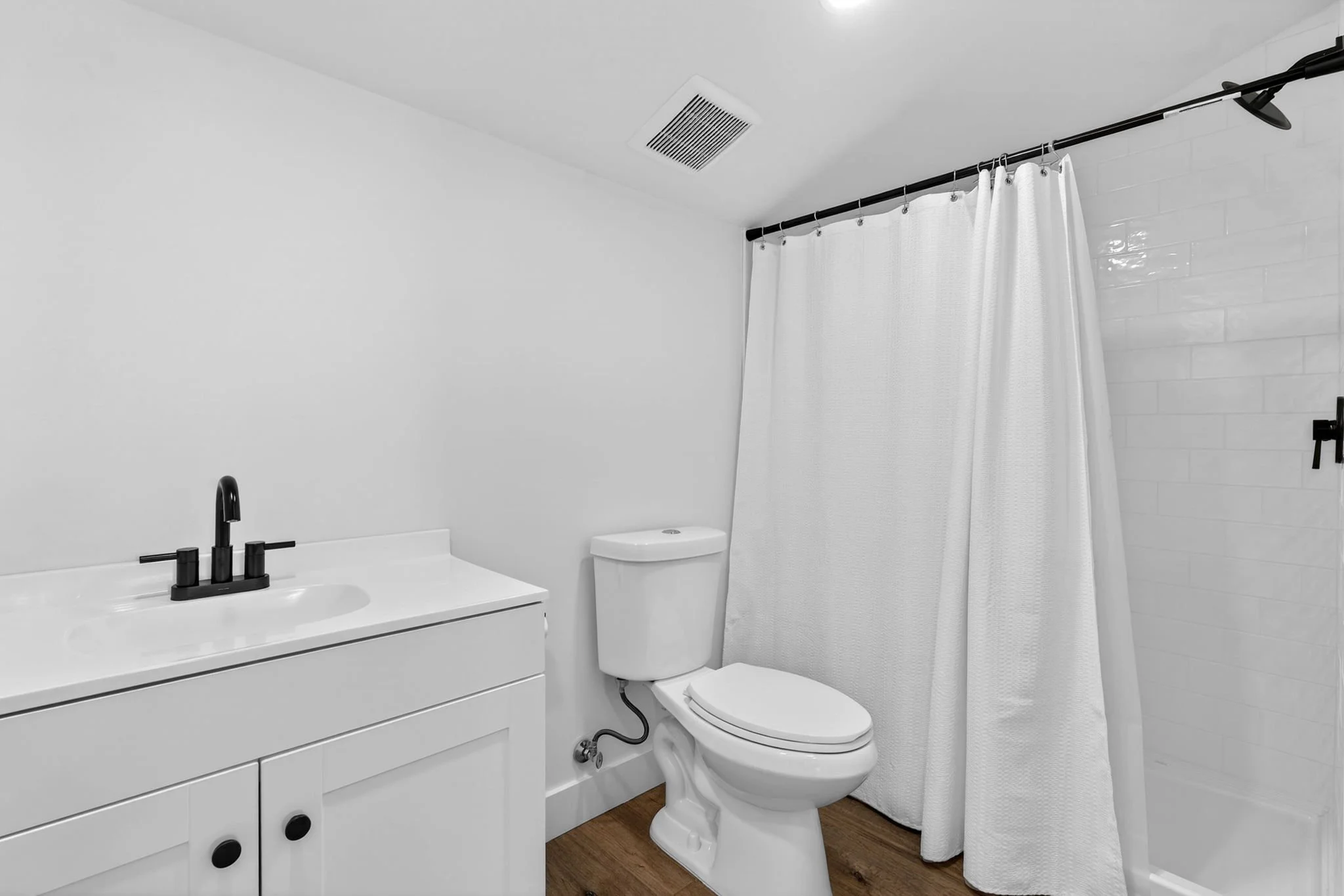 A modern bathroom with a white vanity, black faucet, white toilet, white shower curtain, wall air vent, and wooden flooring.