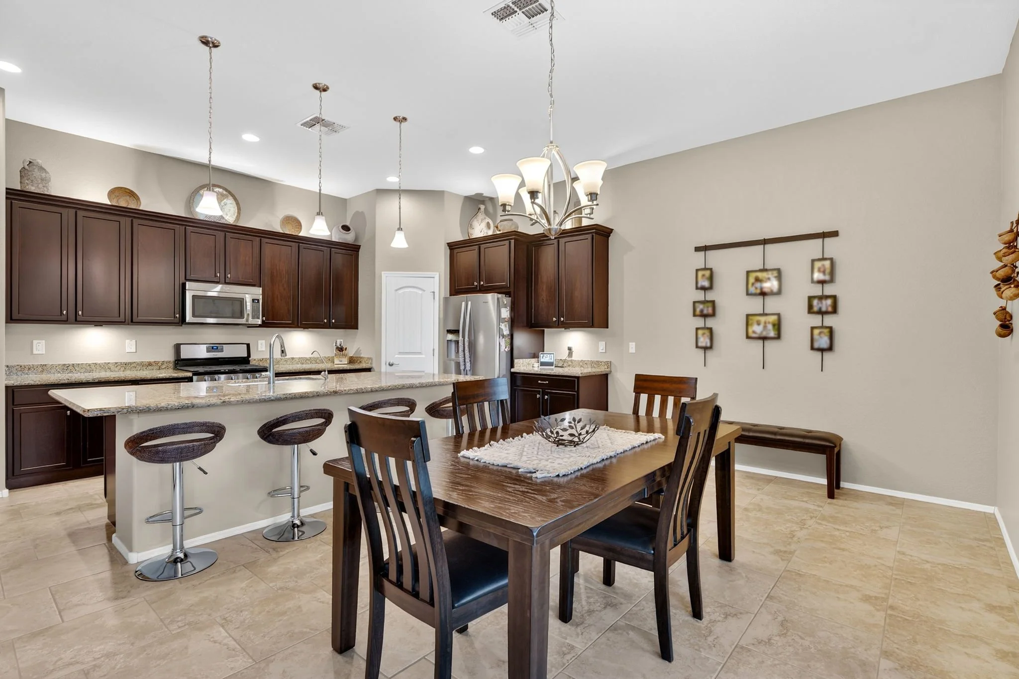 Open-concept kitchen and dining area with dark wood cabinets, stainless steel appliances, granite countertops, a wooden table with six chairs, and decorative pottery on a high shelf. Pendant lights and a chandelier illuminate the space, with framed p