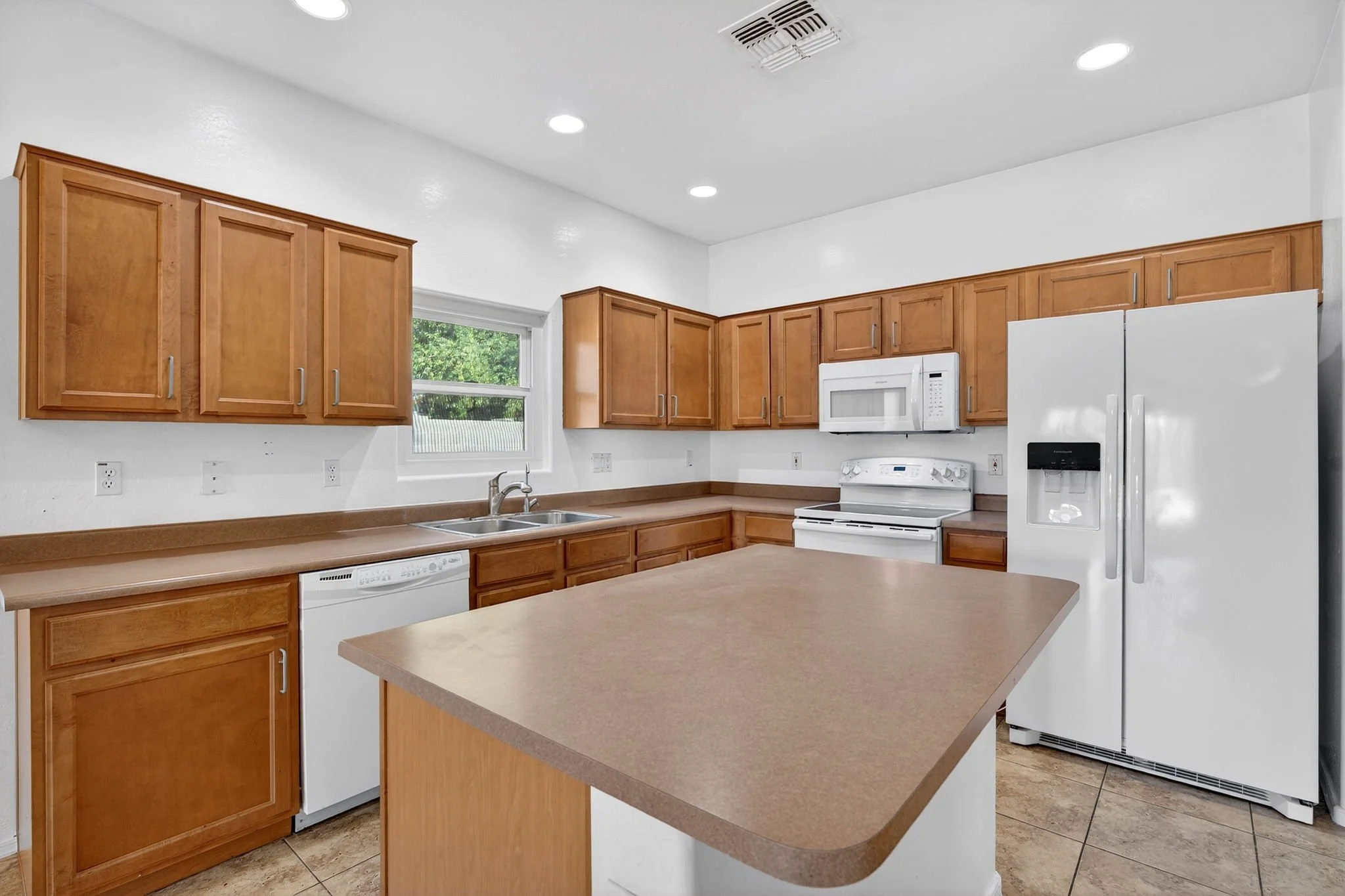 Kitchen with wooden cabinets, white appliances including refrigerator, microwave, and stove, beige countertops, window with green trees outside, ceiling lights, and tiled floor.