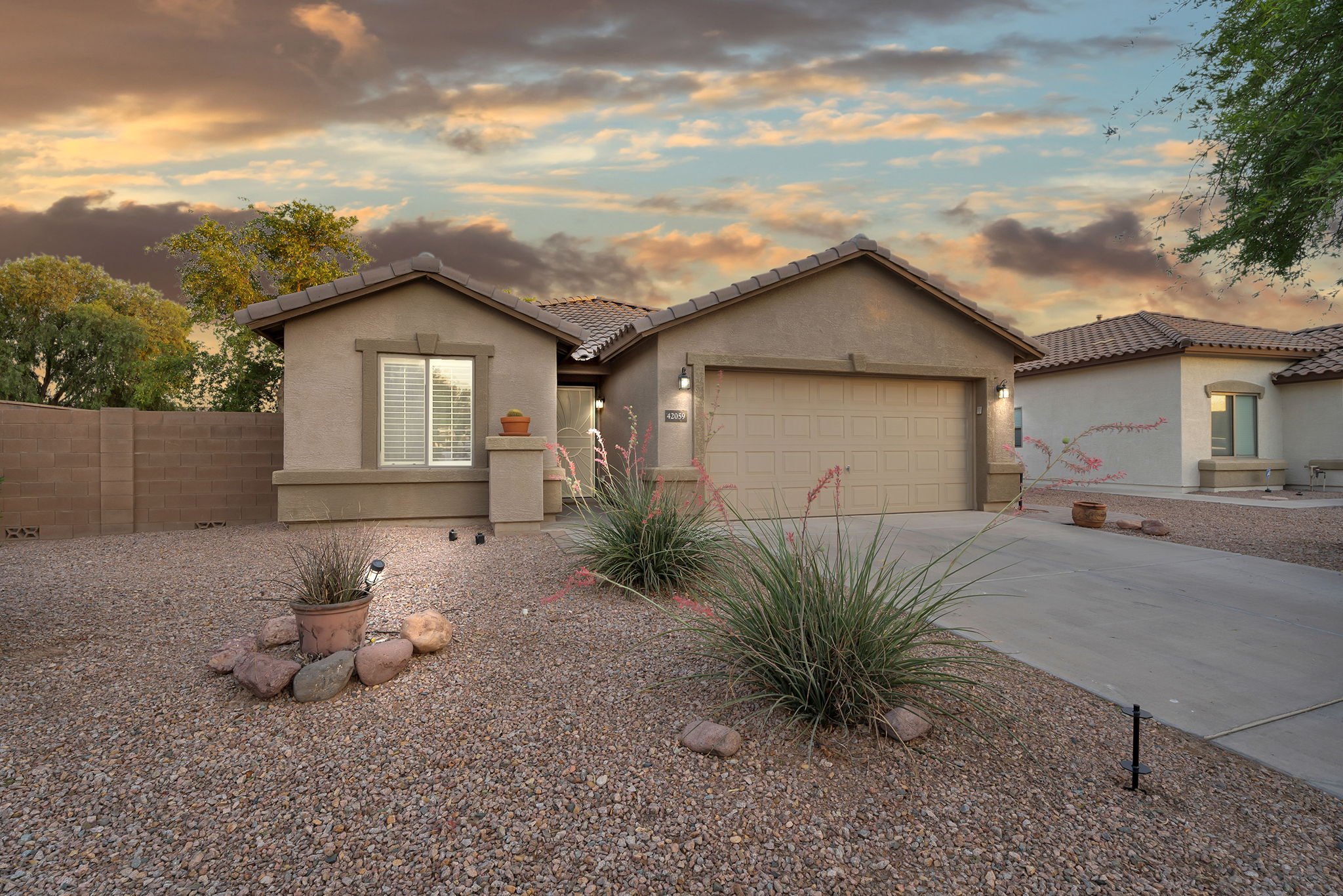 Front yard of a suburban house with desert landscaping, including gravel, rocks, drought-tolerant plants, a potted plant, and small lighting fixtures. The house has a beige stucco exterior, a tiled roof, a garage door, and outdoor lighting. Trees are