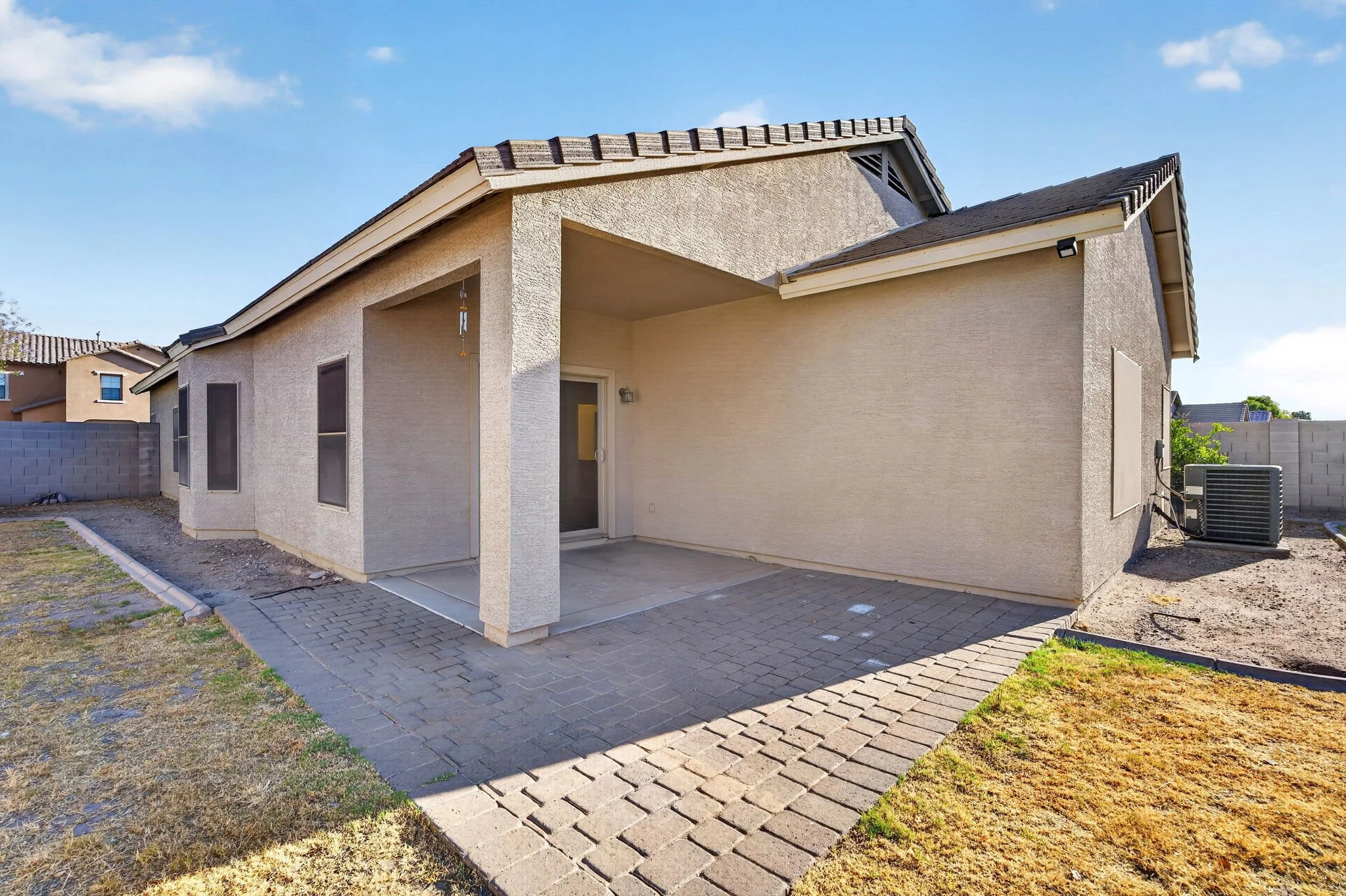 Backyard view of a two-story house with beige stucco walls, a covered patio, and gray paver flooring. There is a small yard with patchy grass and a concrete block wall enclosing the space. An outdoor air conditioning unit is visible to the right, and