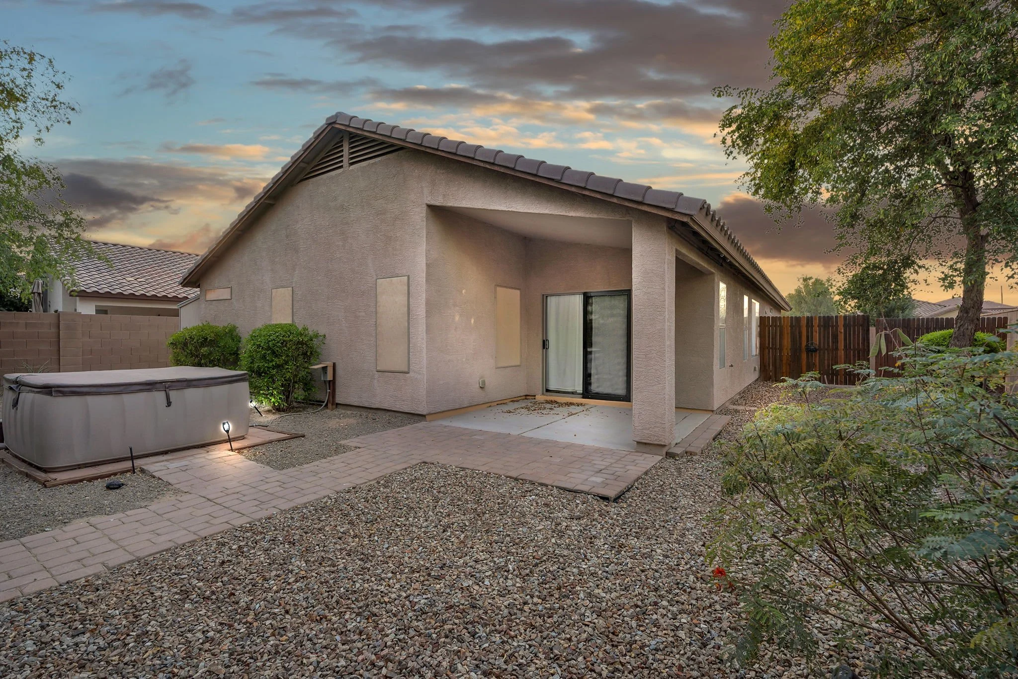 Backyard patio with pavers and gravel, beige house with sliding glass door, trees, and landscaped bushes at sunset.