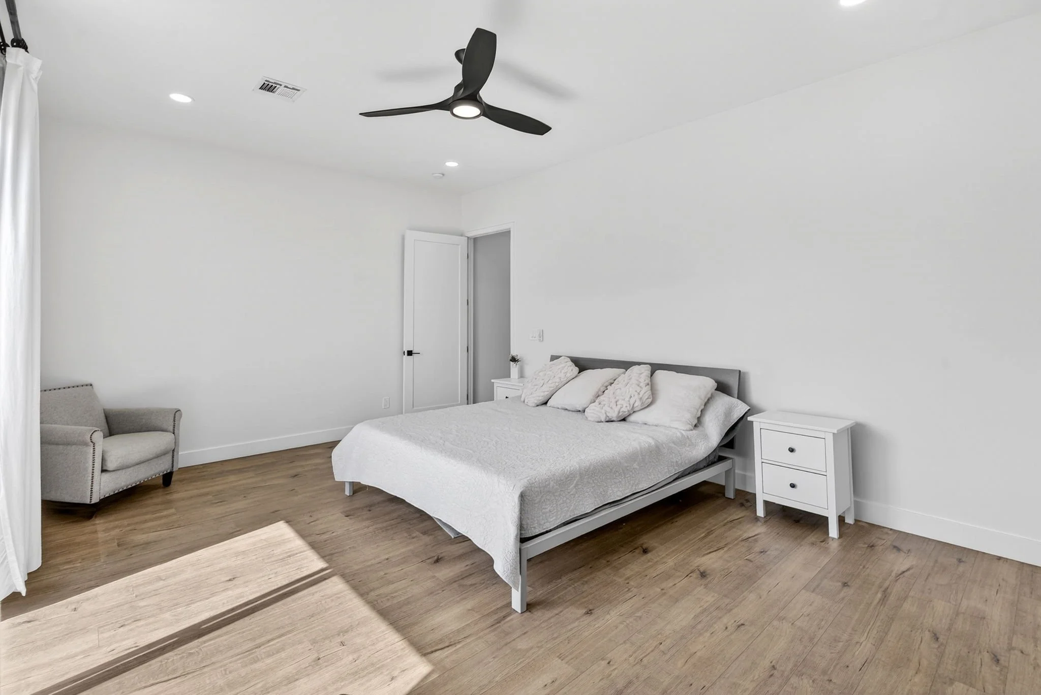 A bright bedroom with a white ceiling, a black ceiling fan, a white bed with pillows, a white nightstand, a gray armchair, and wooden flooring.