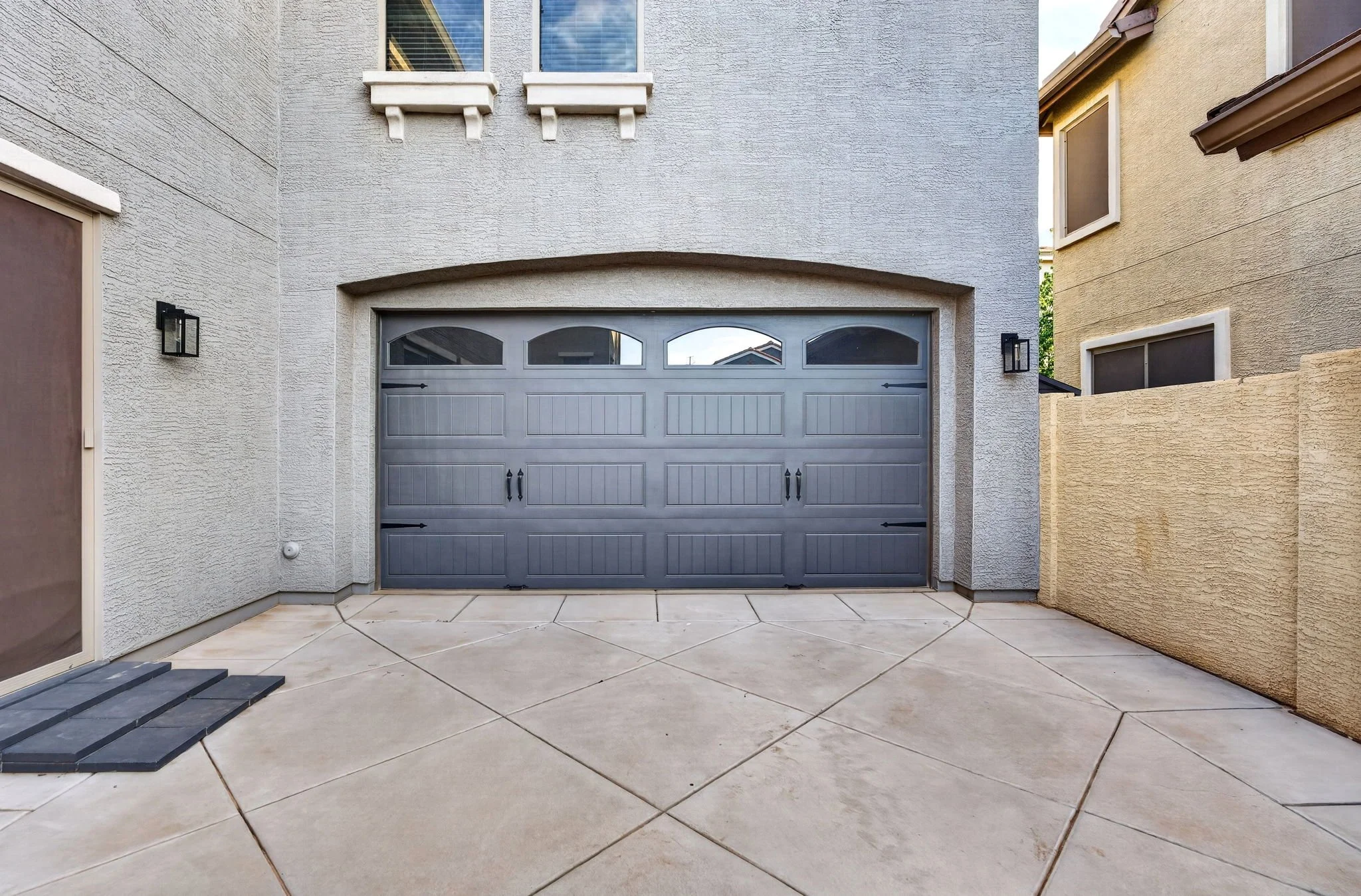 The image shows a gray garage door on a beige stucco house, with beige tile flooring in the foreground and two black wall-mounted lights on either side of the door. There are neighboring houses visible in the background.
