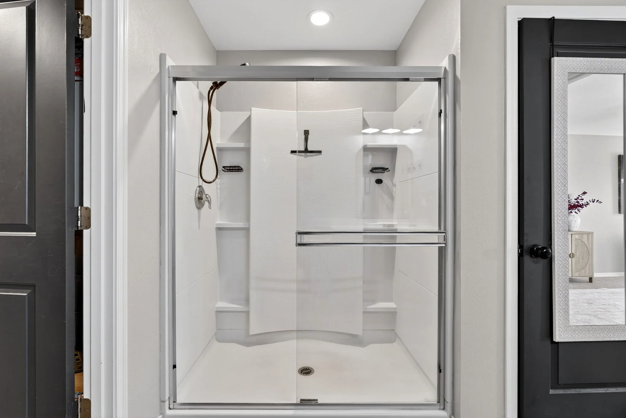 Empty shower with glass door, built-in shelves, handheld showerhead, and a drain, in a modern bathroom.