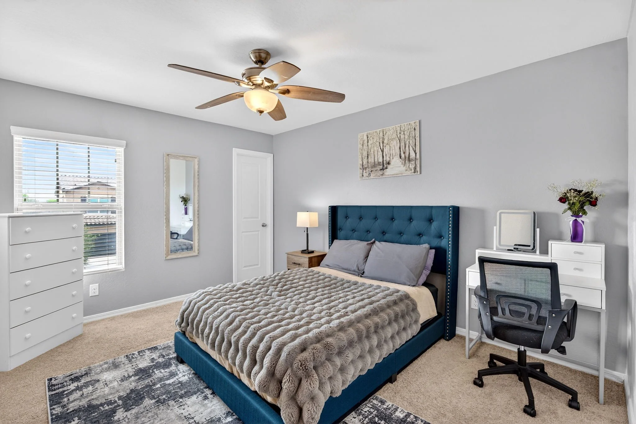 A bedroom with a bed that has a blue frame and gray bedding, a nightstand with a lamp, and a blue headboard. There is a white dresser, a mirror, a window with blinds, and a white desk with a black office chair. The room has light gray walls, a ceilin