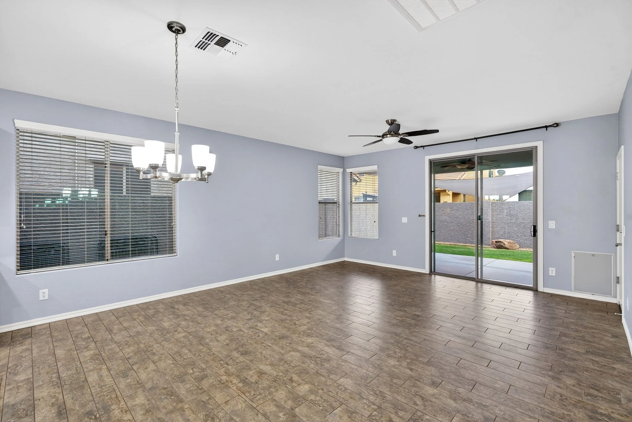 Empty living room with large windows, sliding glass door, ceiling fan, light fixture, light gray walls, brown wooden flooring, and partially visible backyard outside.