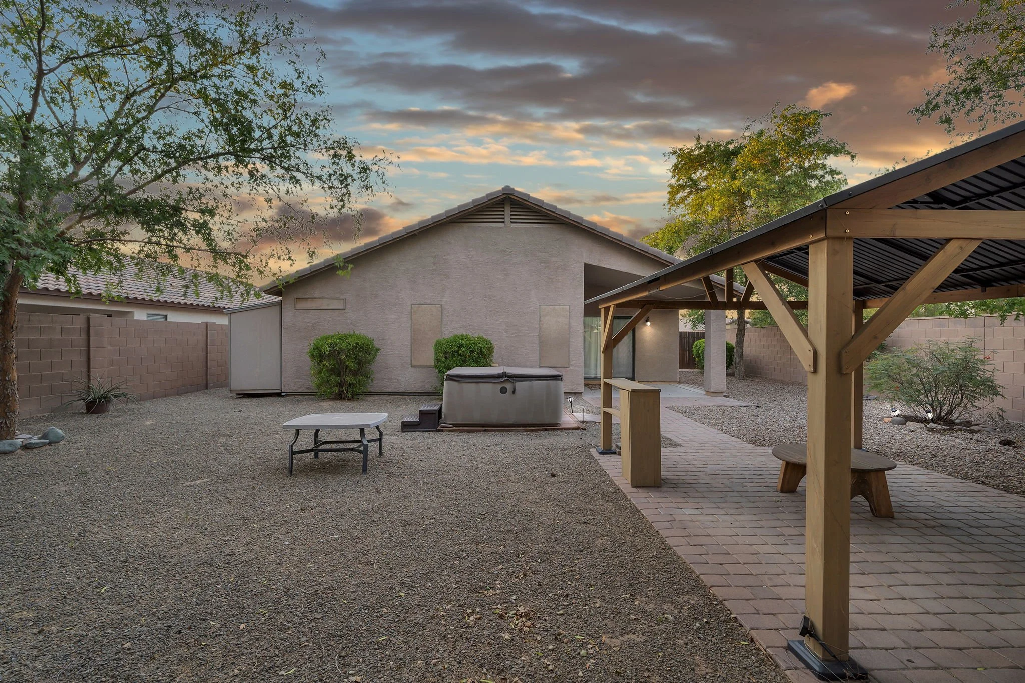 Backyard with gravel ground, a wooden pergola with a metal roof on the right, a small shed, bushes, trees, and a sky with clouds during sunset.