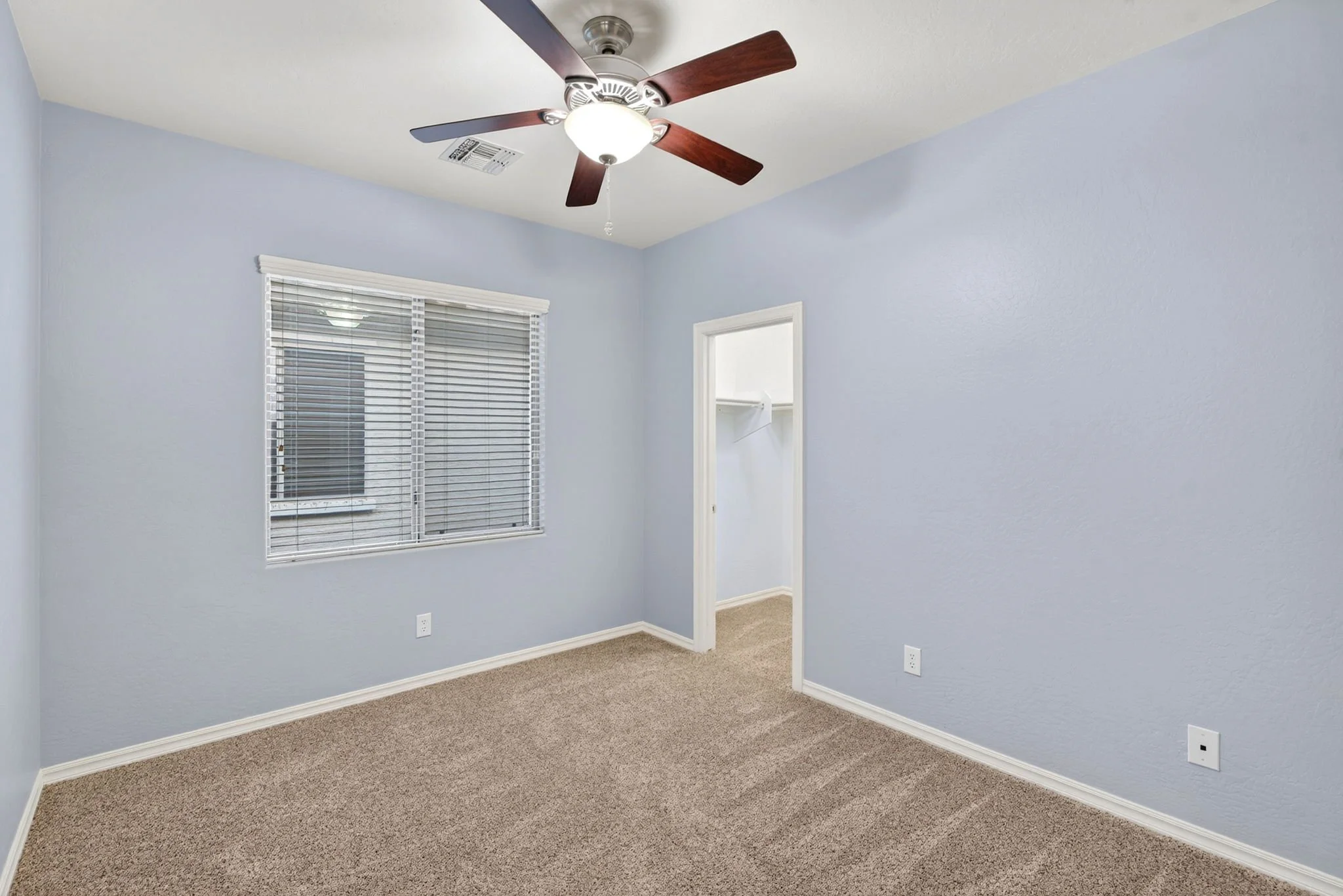 Empty room with light blue walls, a window with blinds, and a ceiling fan with wooden blades.