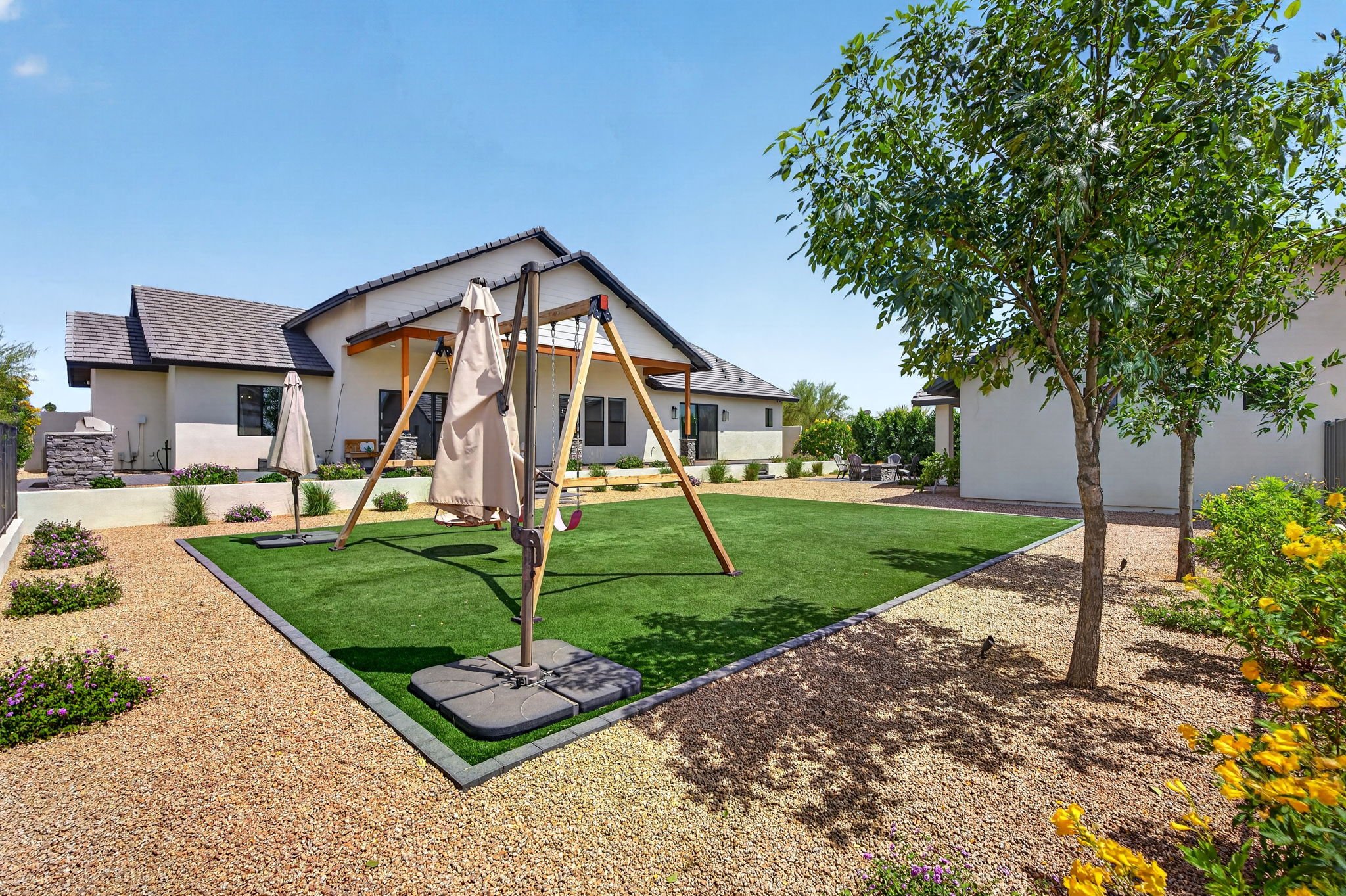 A backyard with a swing set on artificial grass, surrounded by gravel, plants, and trees, with a house in the background under a clear blue sky.