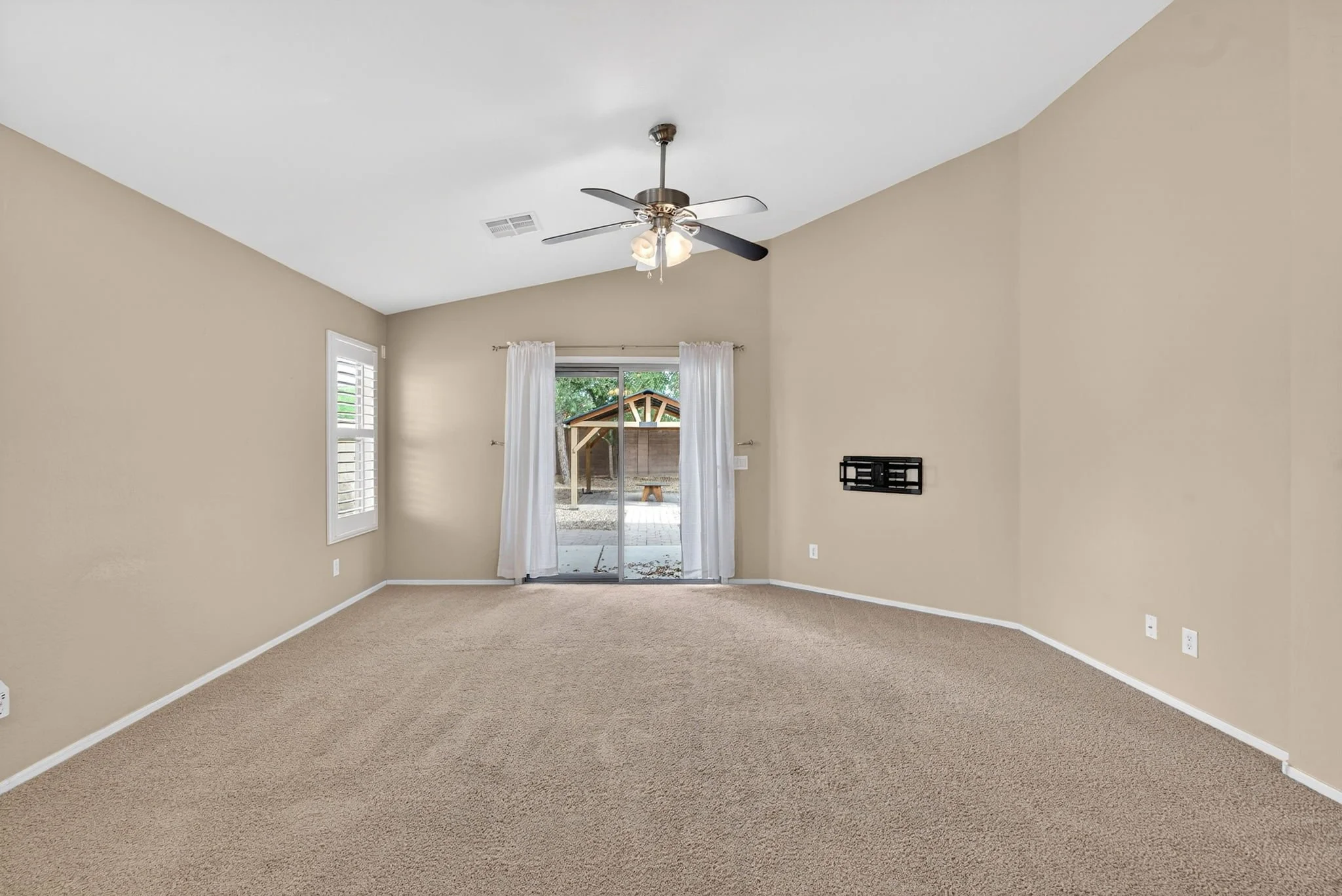 Empty living room with beige walls, brown carpet, windows with white shutters, a sliding glass door with white curtains leading to a patio, a ceiling fan with light, and a wall-mounted TV bracket.