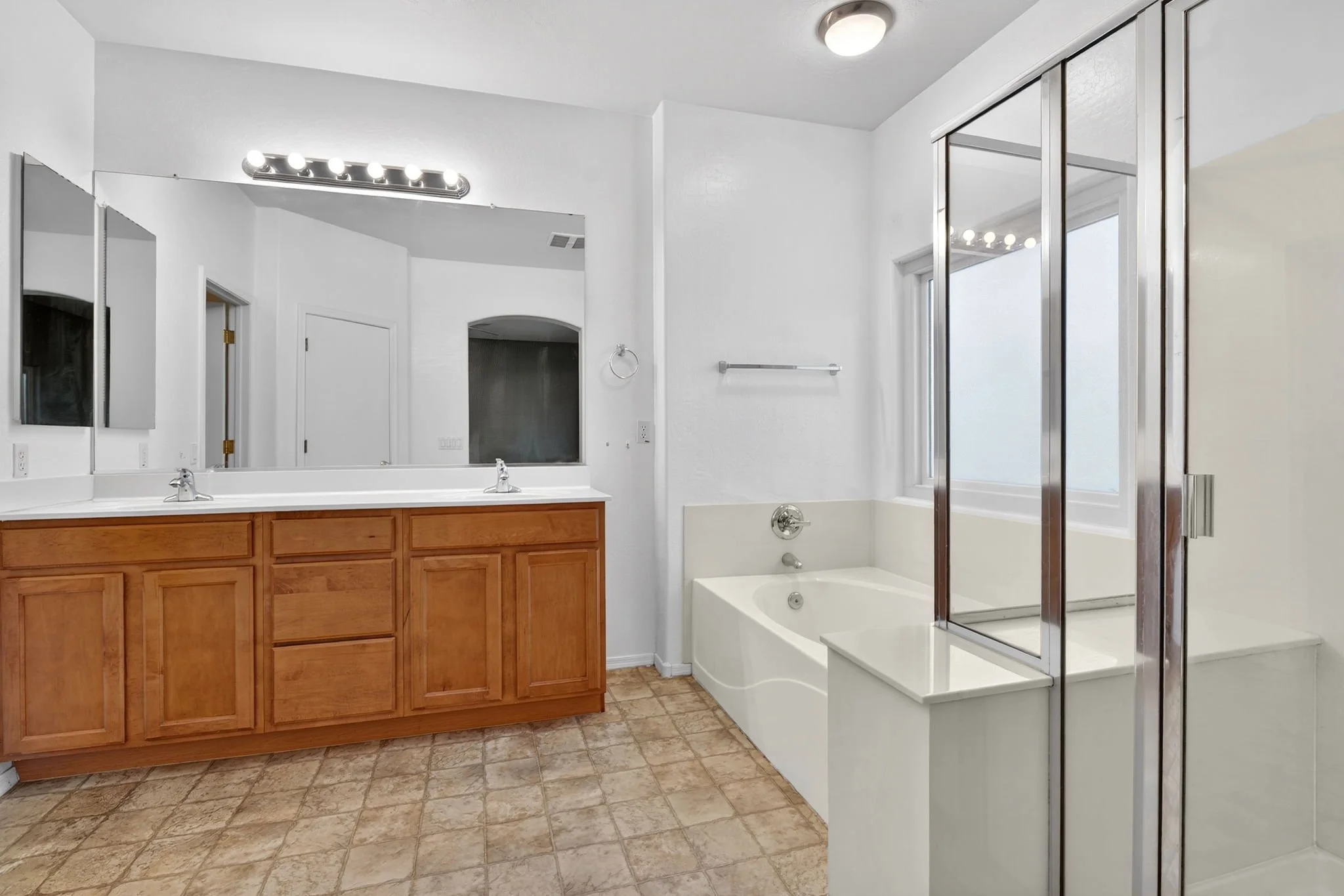 Bathroom with a double vanity, large mirror, vanity lights, bathtub, and shower with glass enclosure, white walls, and tiled floor.