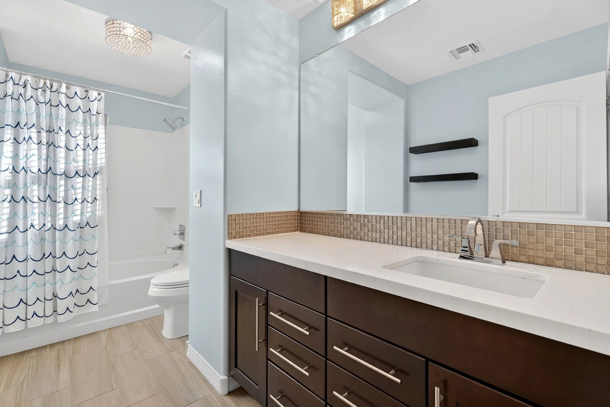 Modern bathroom with a shower area separated by a low wall, a vanity with dark wood drawers, a large mirror, black floating shelves, beige tile accents, and a light blue wall color.
