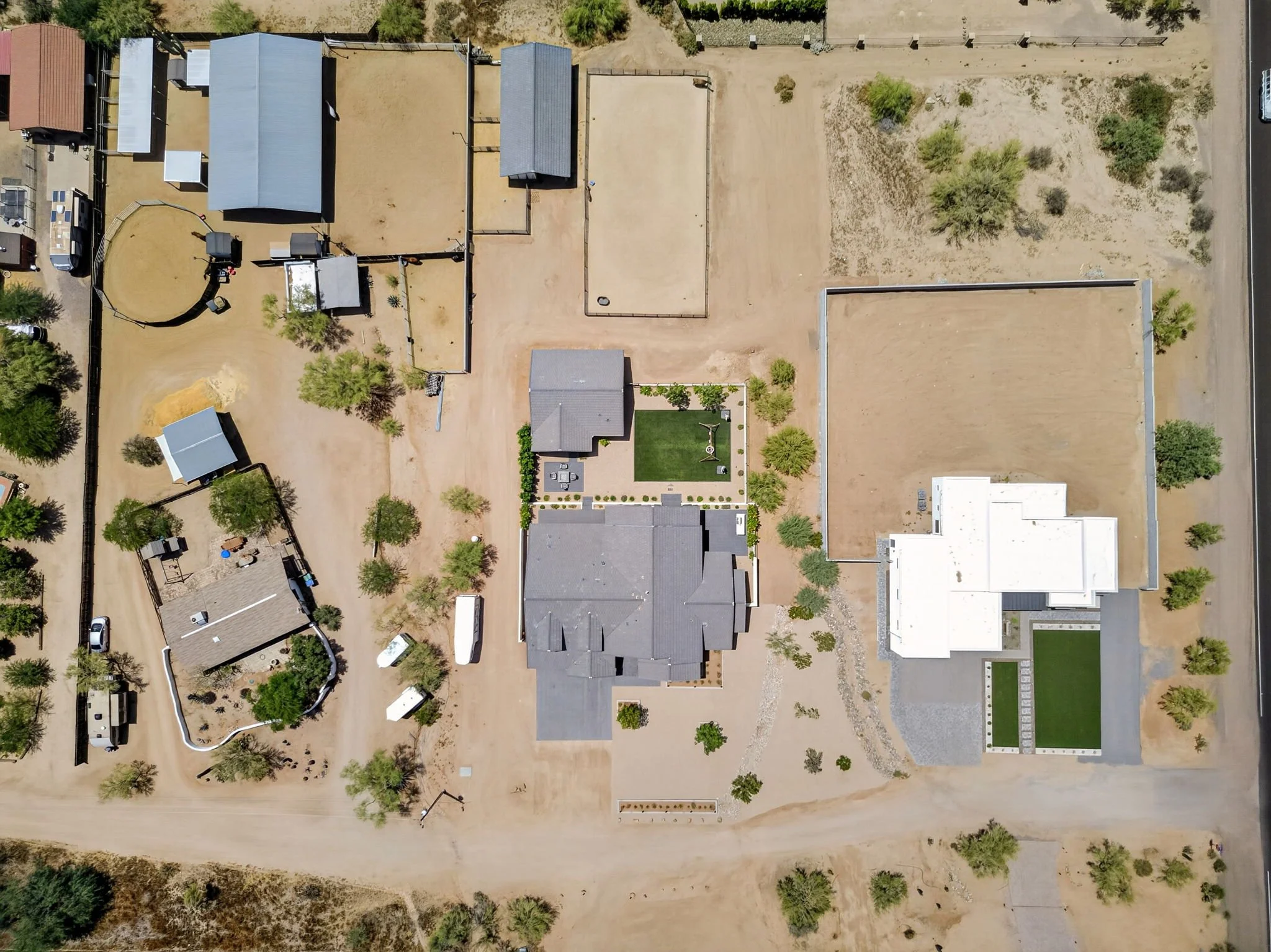 Aerial view of a residential backyard yard with a sandy and dirt lawn, several trees, and fences. There are multiple houses, some with patios and garden areas, and a few sports courts or open sandy areas.