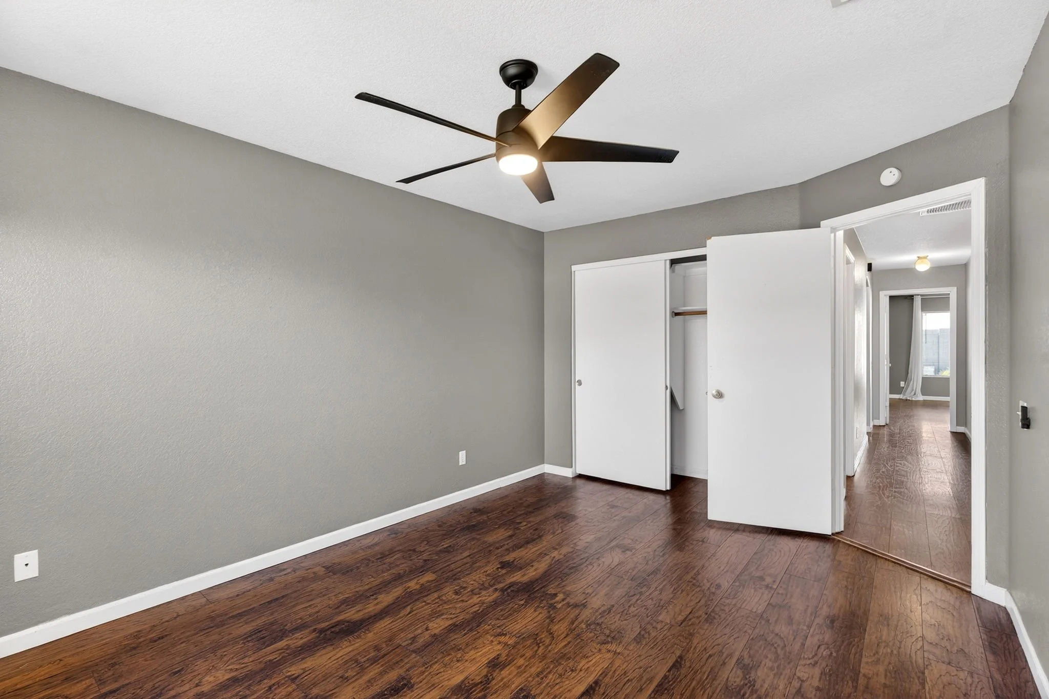 Empty room with gray walls, dark hardwood floor, ceiling fan, closet door partially open, and hallway leading to other rooms.