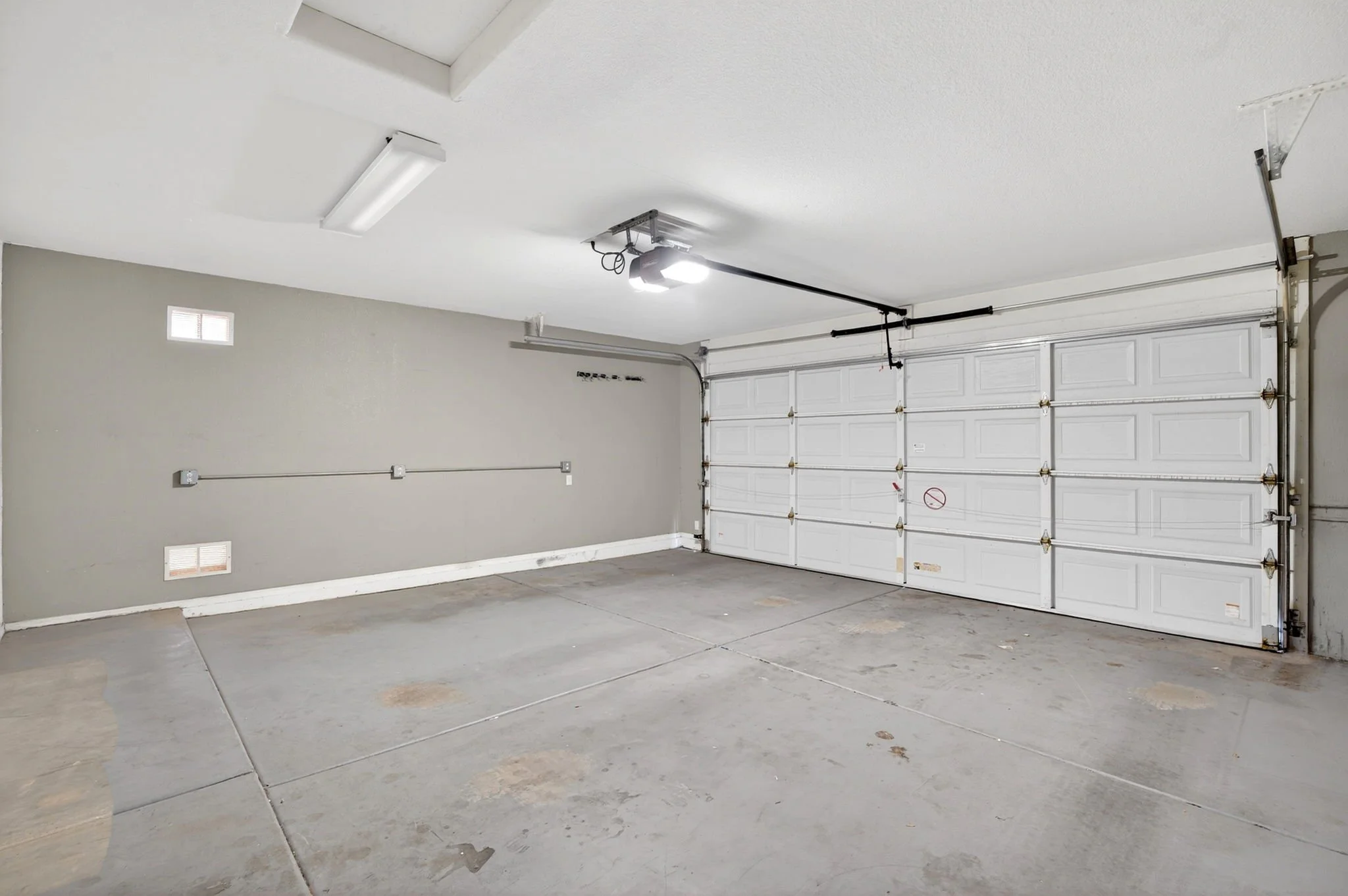 Empty two-car garage with closed white garage door, concrete floor, gray walls, garage door opener, and ceiling light.
