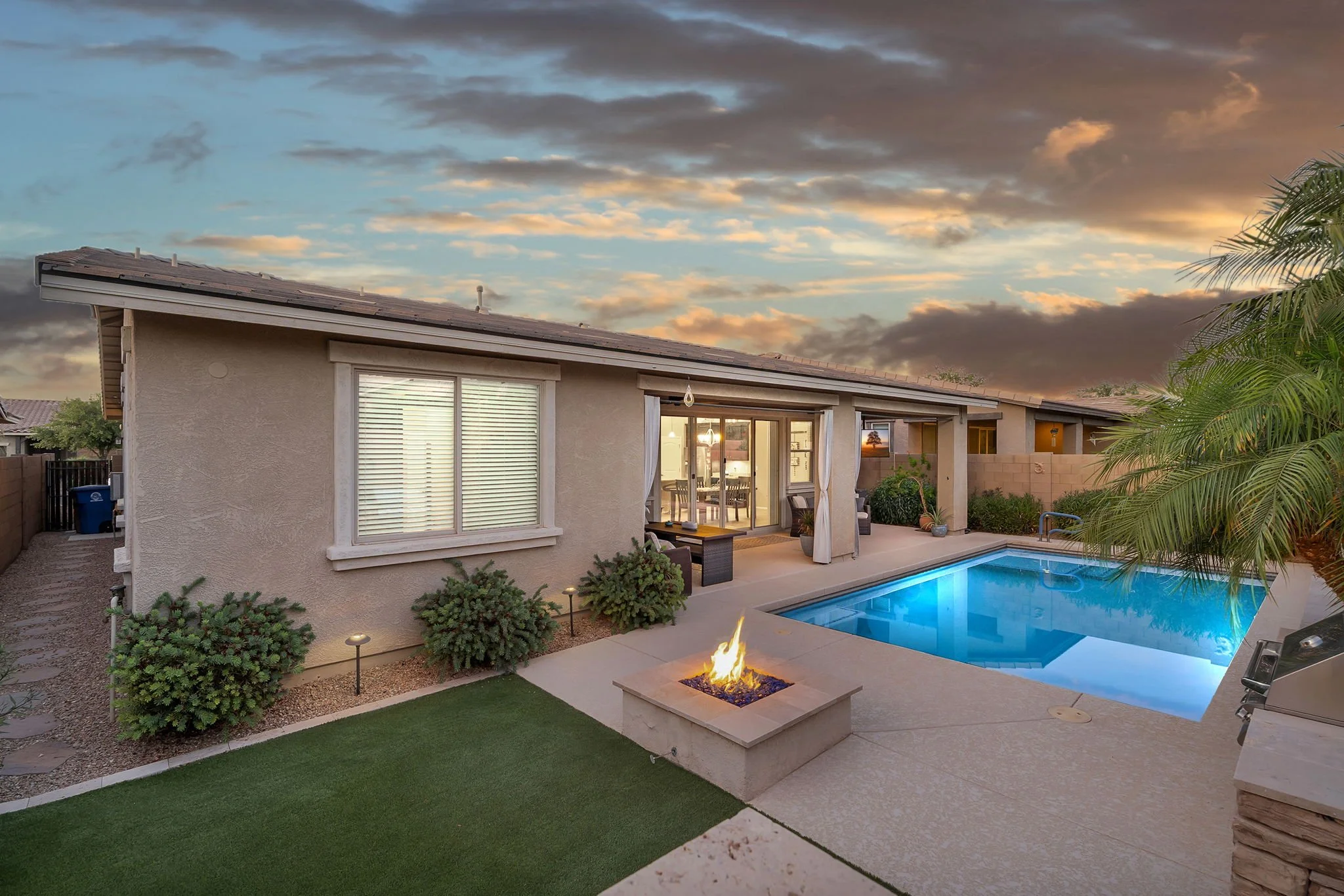 A suburban backyard featuring a swimming pool with blue water, a stone fire pit with flames, and a patio area with outdoor furniture. The house has large sliding glass doors leading to the interior, and there are plants and small bushes along the yar
