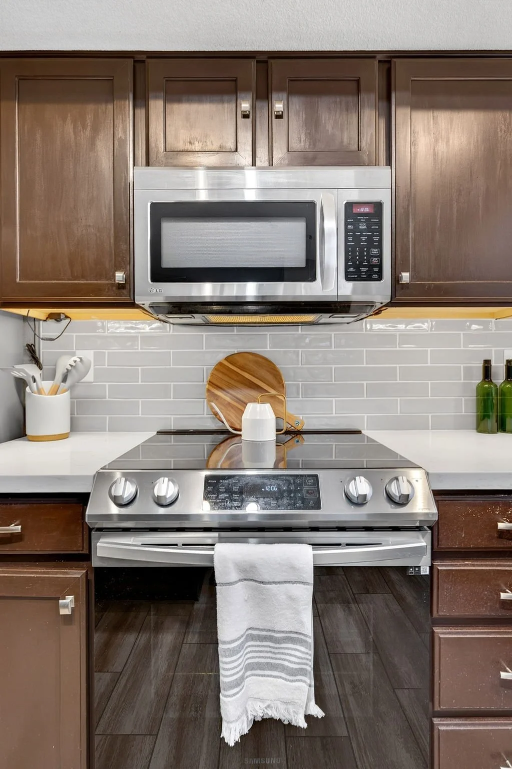 Kitchen with brown cabinets, stainless steel microwave, stove, white countertops, white subway tile backsplash, utensils in a white container, cutting board, green bottles, and a white towel hanging from oven handle.