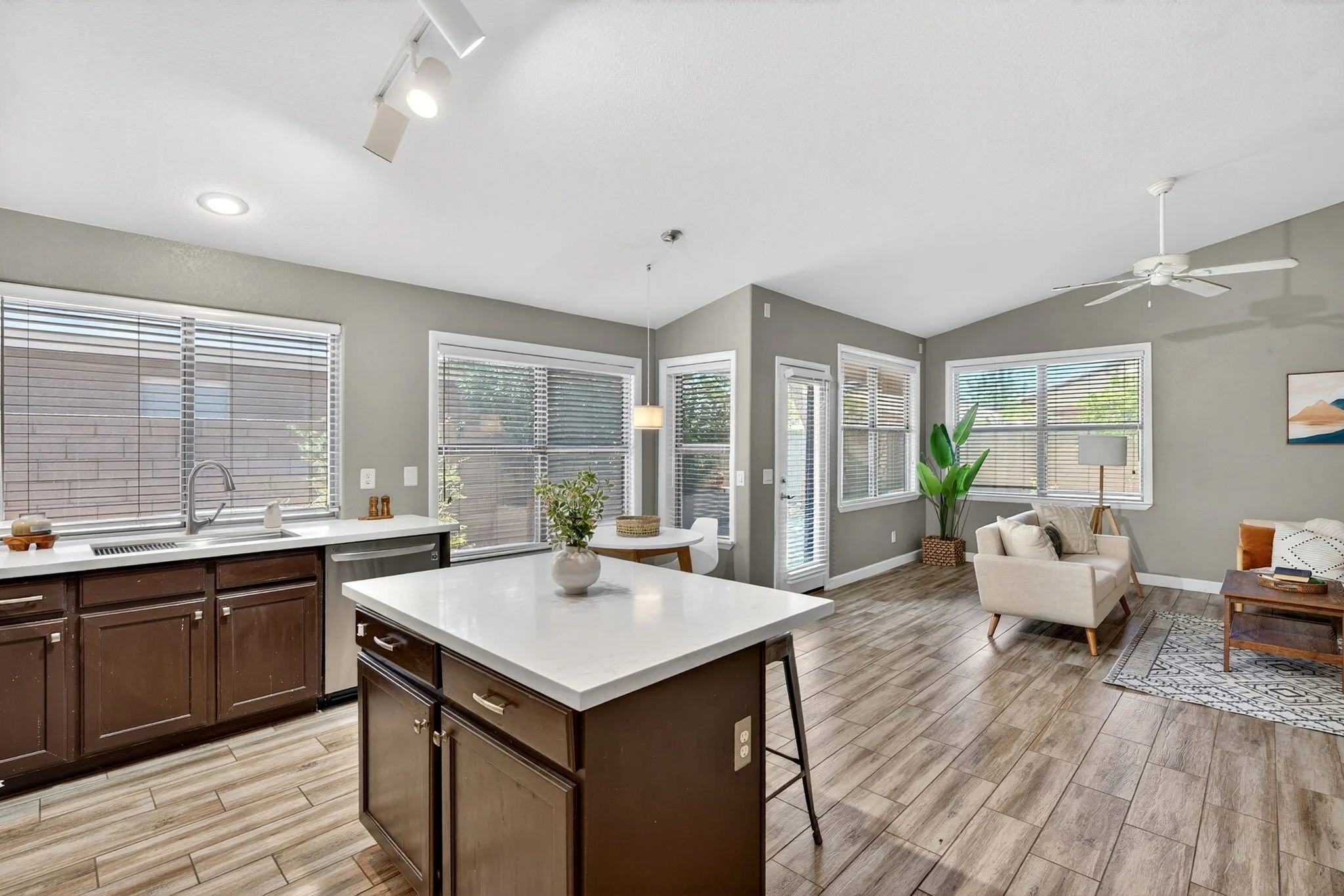 Open-concept kitchen and living room with large windows, white kitchen island, dark wood cabinets, beige sofa, potted plant, and hardwood flooring.