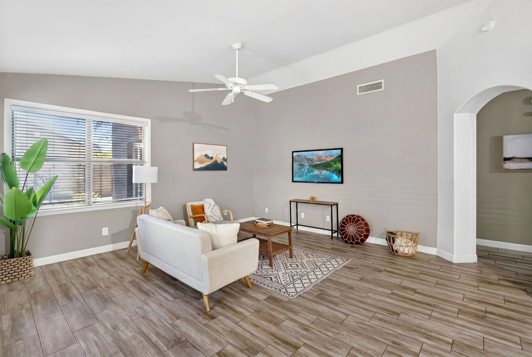 Living room with a white ceiling fan, large window with blinds, gray walls, modern art, small black TV stand with a landscape image on the TV, white sofa, wooden armchairs, patterned rug, potted plant, baskets, and wood flooring.
