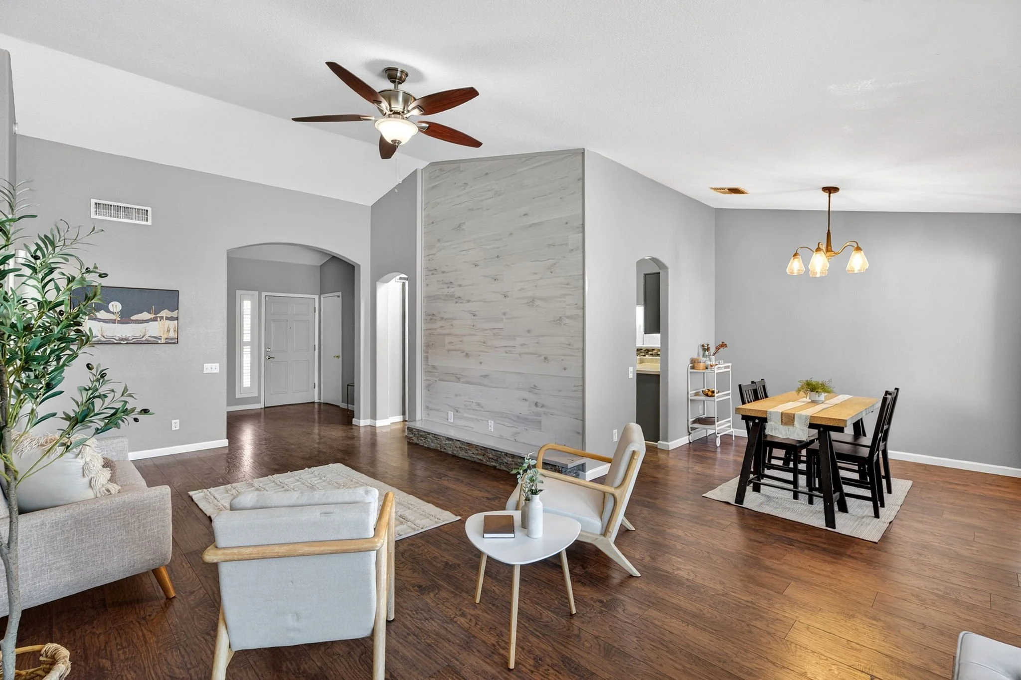 Open living room with gray walls, two beige armchairs, a white small round table with a book and a vase, a wooden dining table with chairs, a ceiling fan, and a chandelier over the dining area.