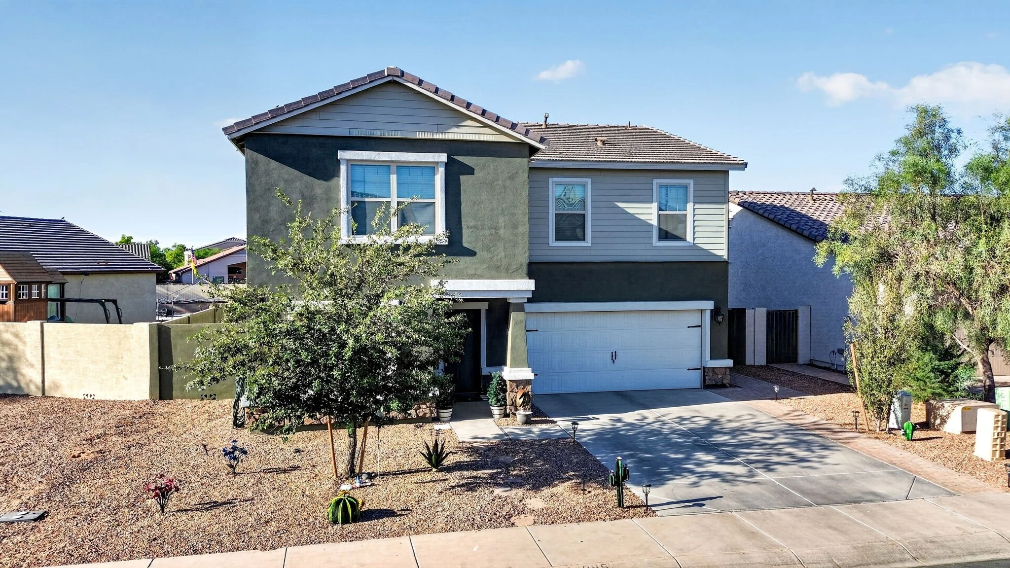 Front view of a modern two-story house with a grey and white exterior, a two-car garage, and a landscaped yard with trees and small plants under a clear blue sky.