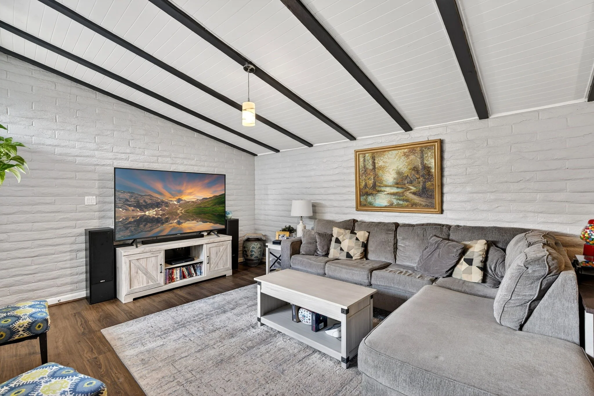 Living room with white brick walls, ceiling beams, a TV on a white stand, a gray sectional sofa with patterned pillows, a framed landscape painting, a white table, a lamp, a potted plant, and a multicolored gumball machine.