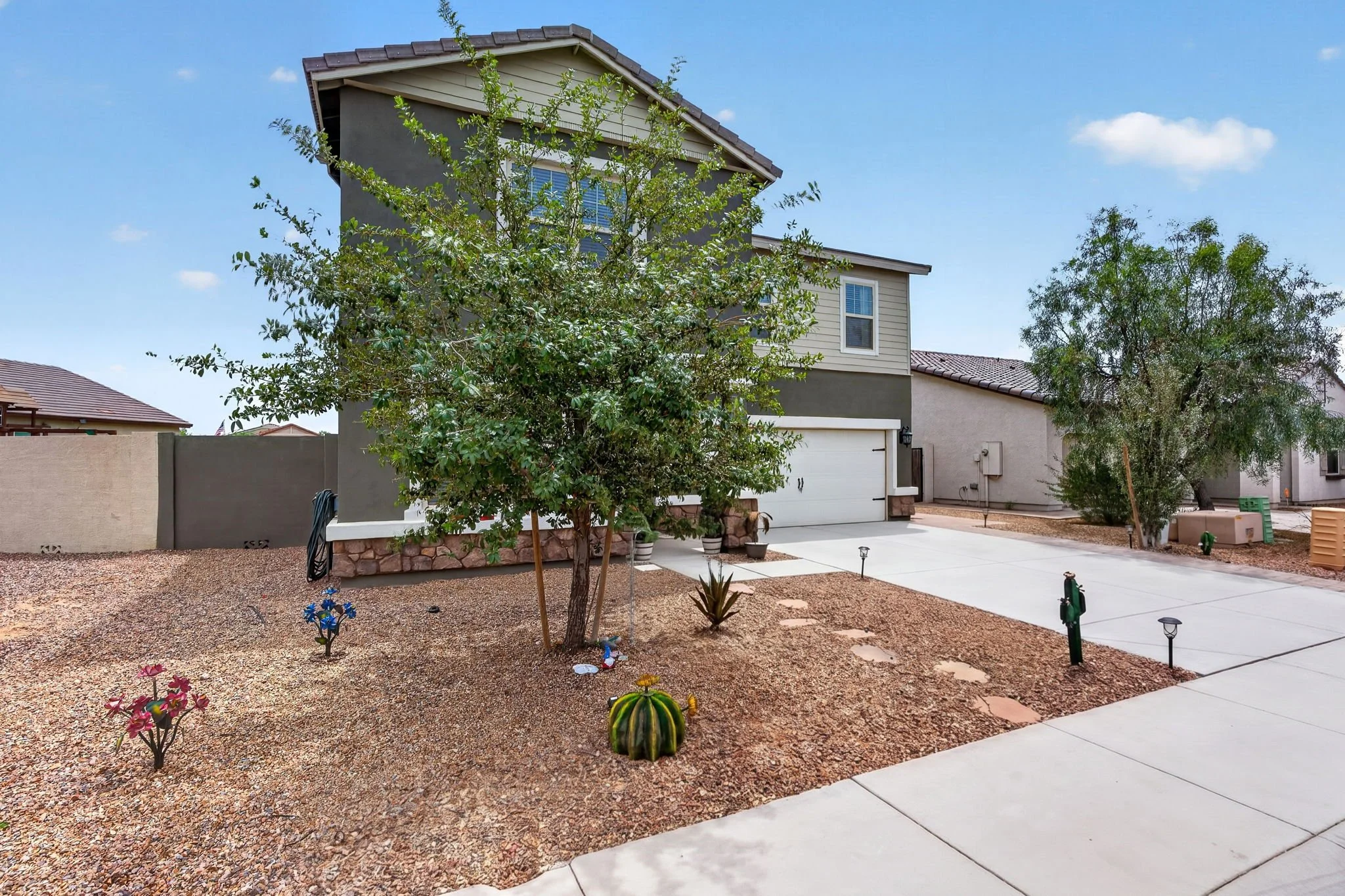 Front yard of a modern two-story house with a concrete driveway, desert landscaping, and various cacti and plants, under a clear blue sky.