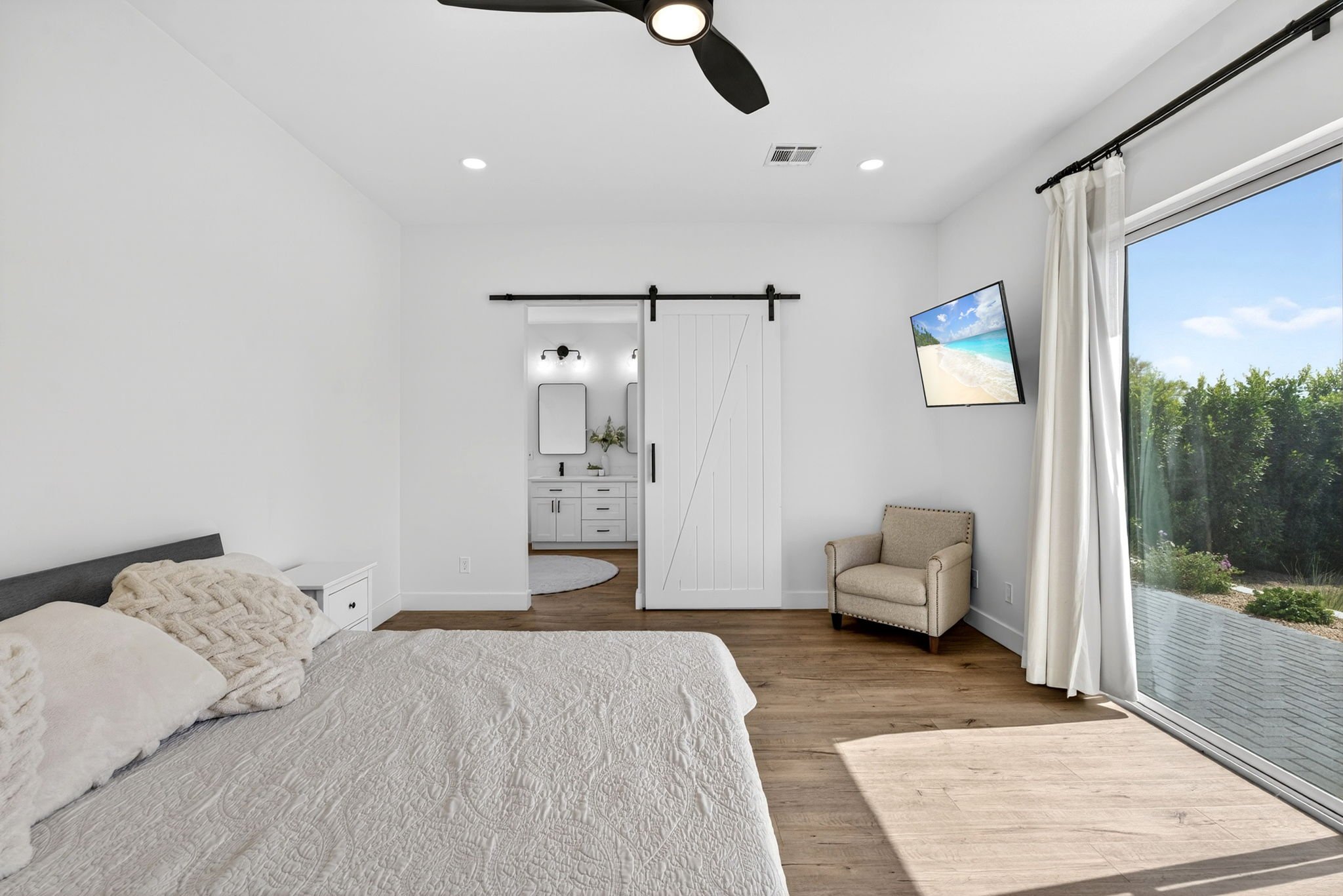 A bedroom with white walls, a bed with textured pink bedding, and pillows. There is a sliding glass door with white curtains leading to an outdoor deck. A beige armchair is near the window. A flat-screen TV displays a beach scene and is mounted on th