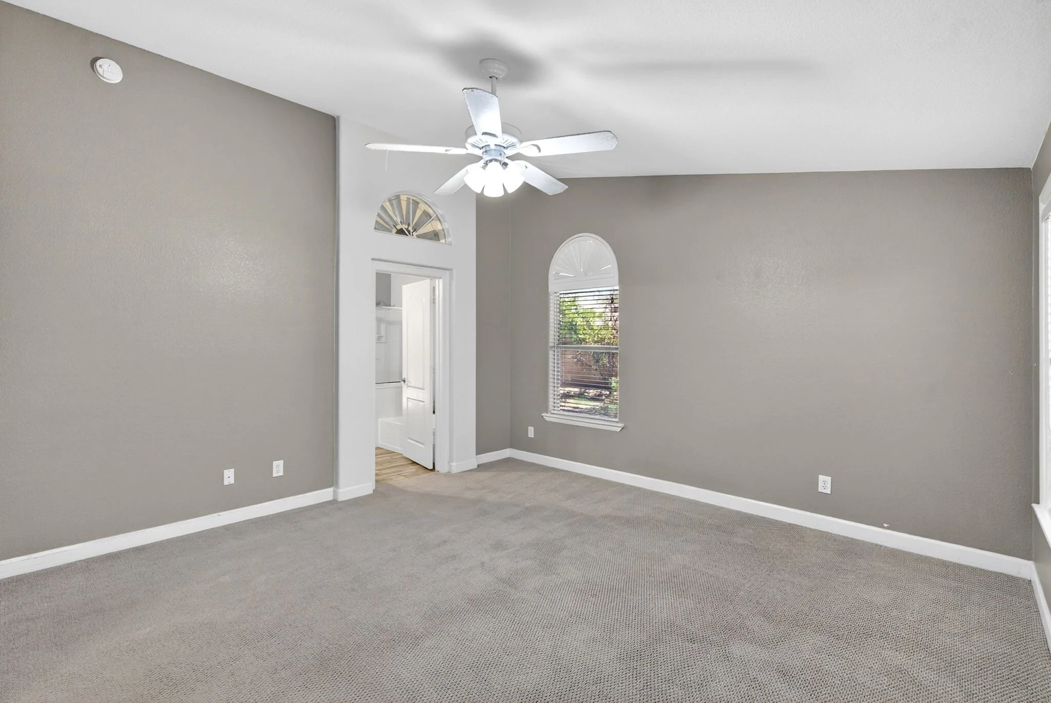 Empty living room with gray walls, white ceiling fan, carpeted floor, and windows with blinds.