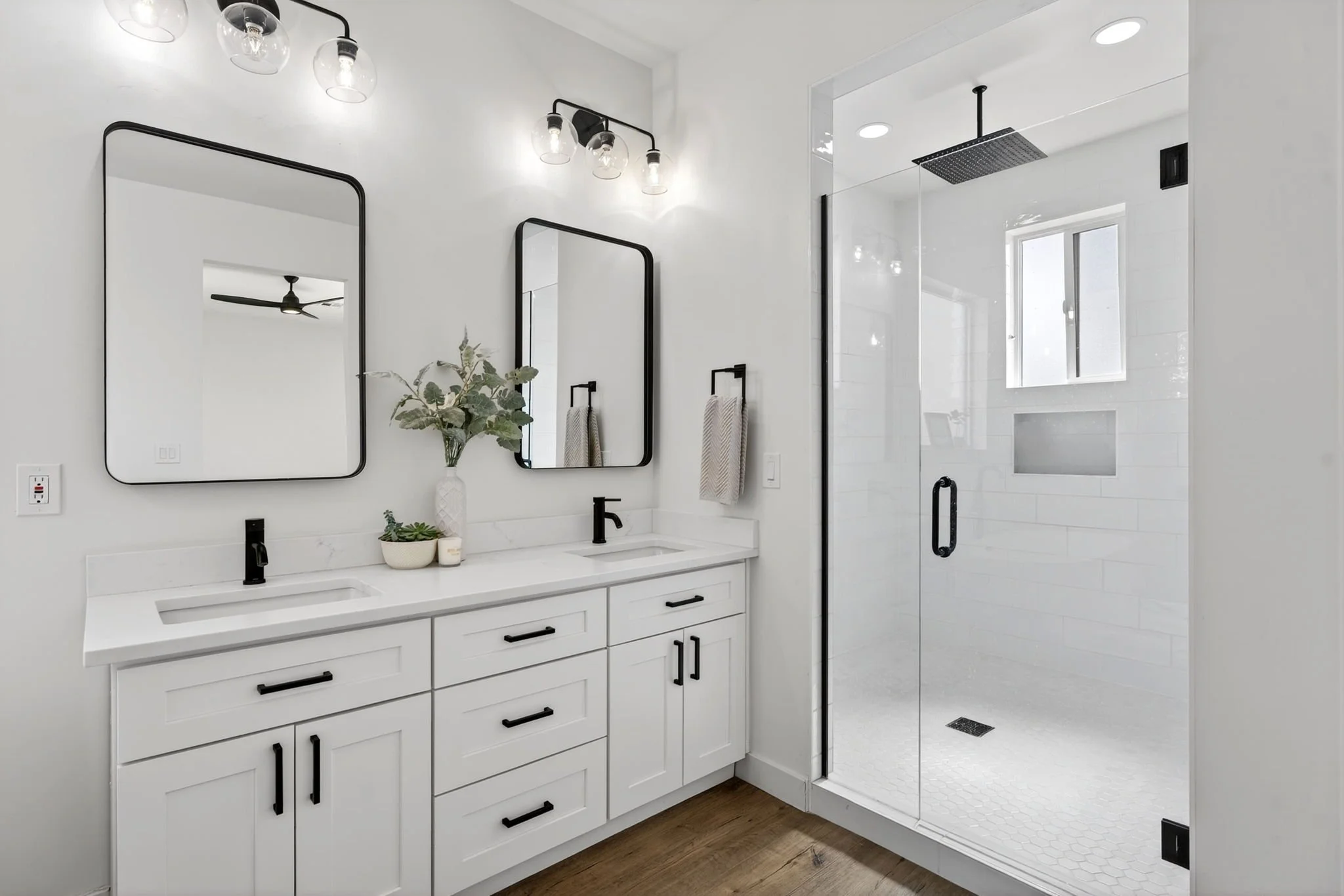 Modern bathroom with white vanity, black hardware, and two black-framed mirrors. There are two black faucets and a vase with green plants on the counter. A glass shower with a rainfall showerhead is visible, with a window providing natural light.