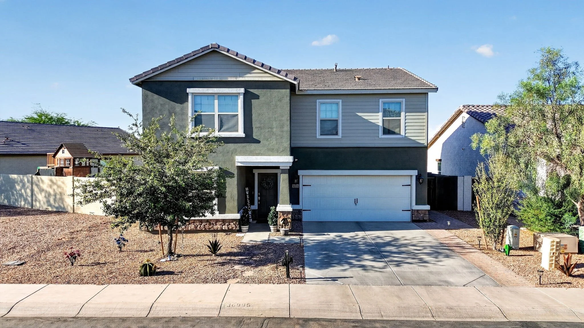 Front view of a modern two-story house with a gray and beige exterior, white trim, a two-car garage, small front yard with desert landscaping, and clear blue sky.