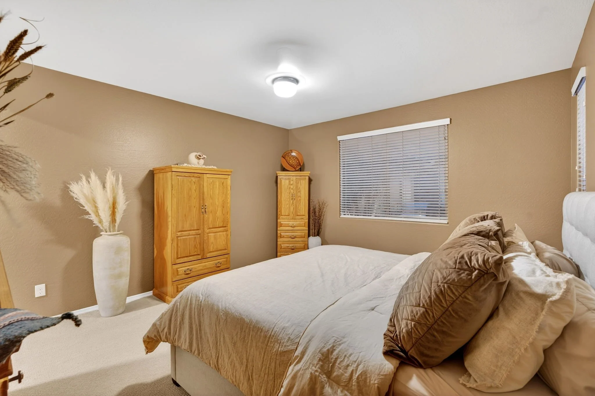 A bedroom with beige walls, a large bed with brown and beige bedding, two wooden dressers, a window with blinds, and decorative items including vases and a spherical sculpture.