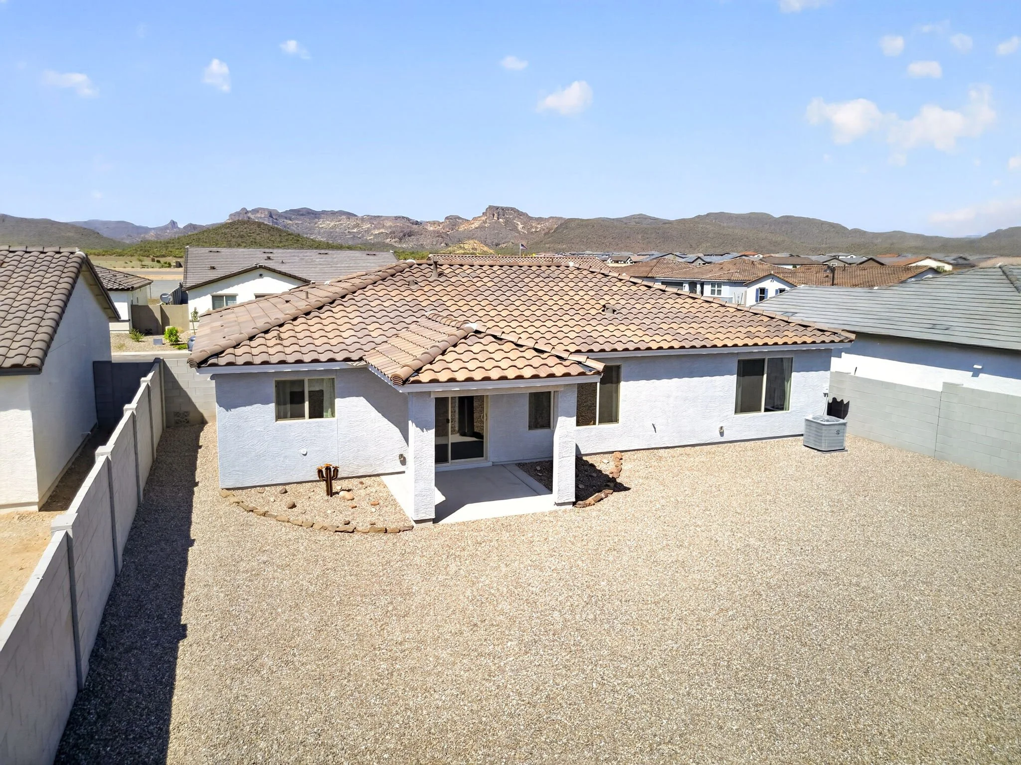 View of a backyard with a gravel ground, white house with a tiled roof, and a mountain range in the background under a blue sky with scattered clouds.