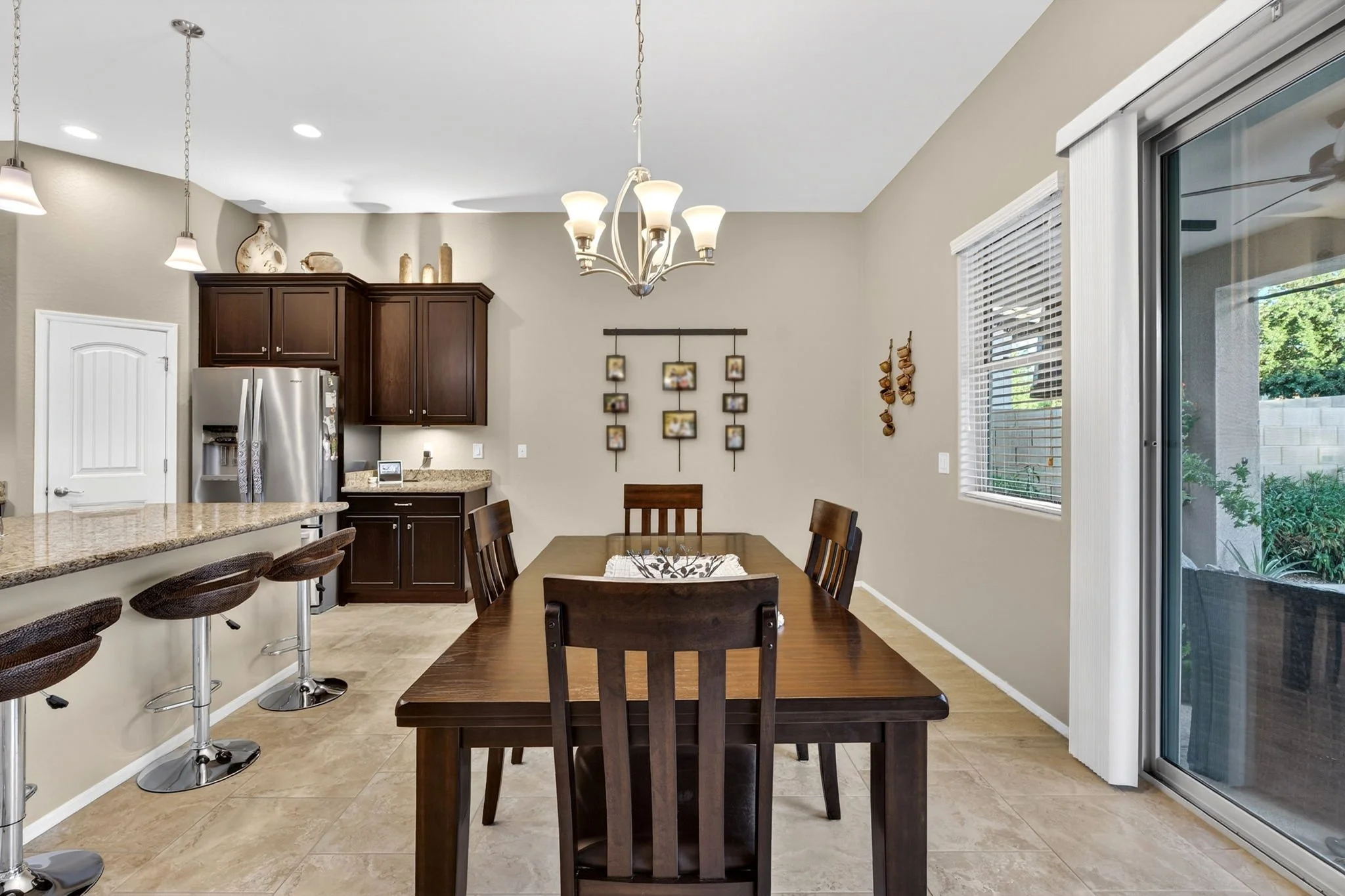 Dining area with a wooden table and chairs, kitchen with dark cabinets, granite counters, and a stainless steel refrigerator, large window with blinds, sliding glass door, chandelier, and decorative items on the walls and shelves.