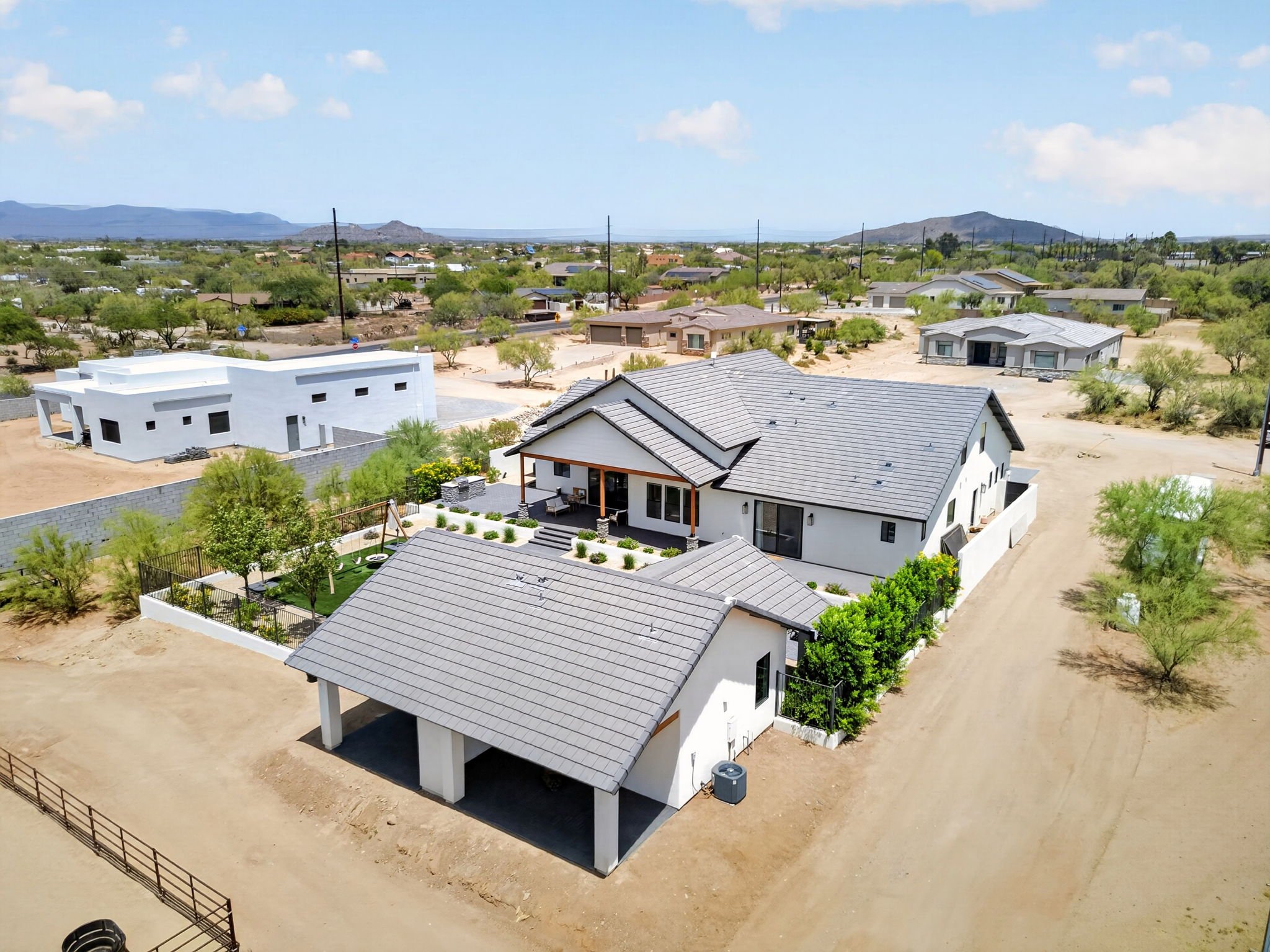 Aerial view of a residential neighborhood featuring newly built modern homes with gray and white exteriors, surrounded by desert landscape and distant mountains under a partly cloudy sky.