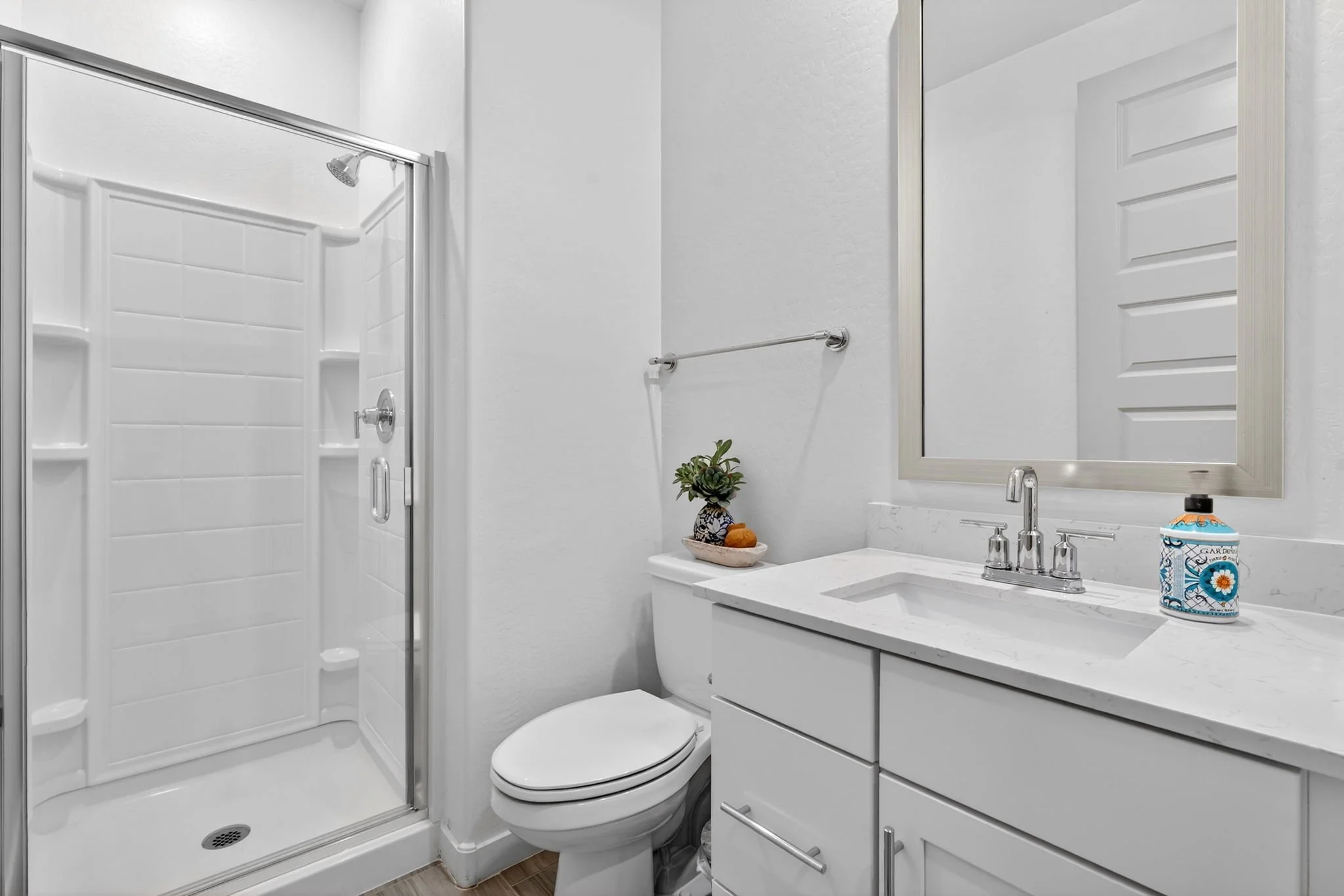 A small, modern bathroom with a white vanity, mirror, and sink, a toilet, and a glass shower with a sliding door. There is a decorative plant and toiletries on the counter.