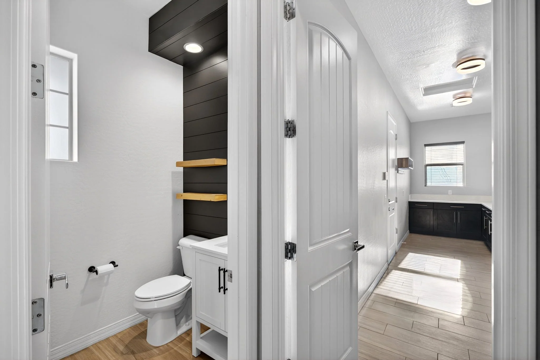 Small bathroom with toilet, white walls, and wood flooring, partially viewable from an open door.