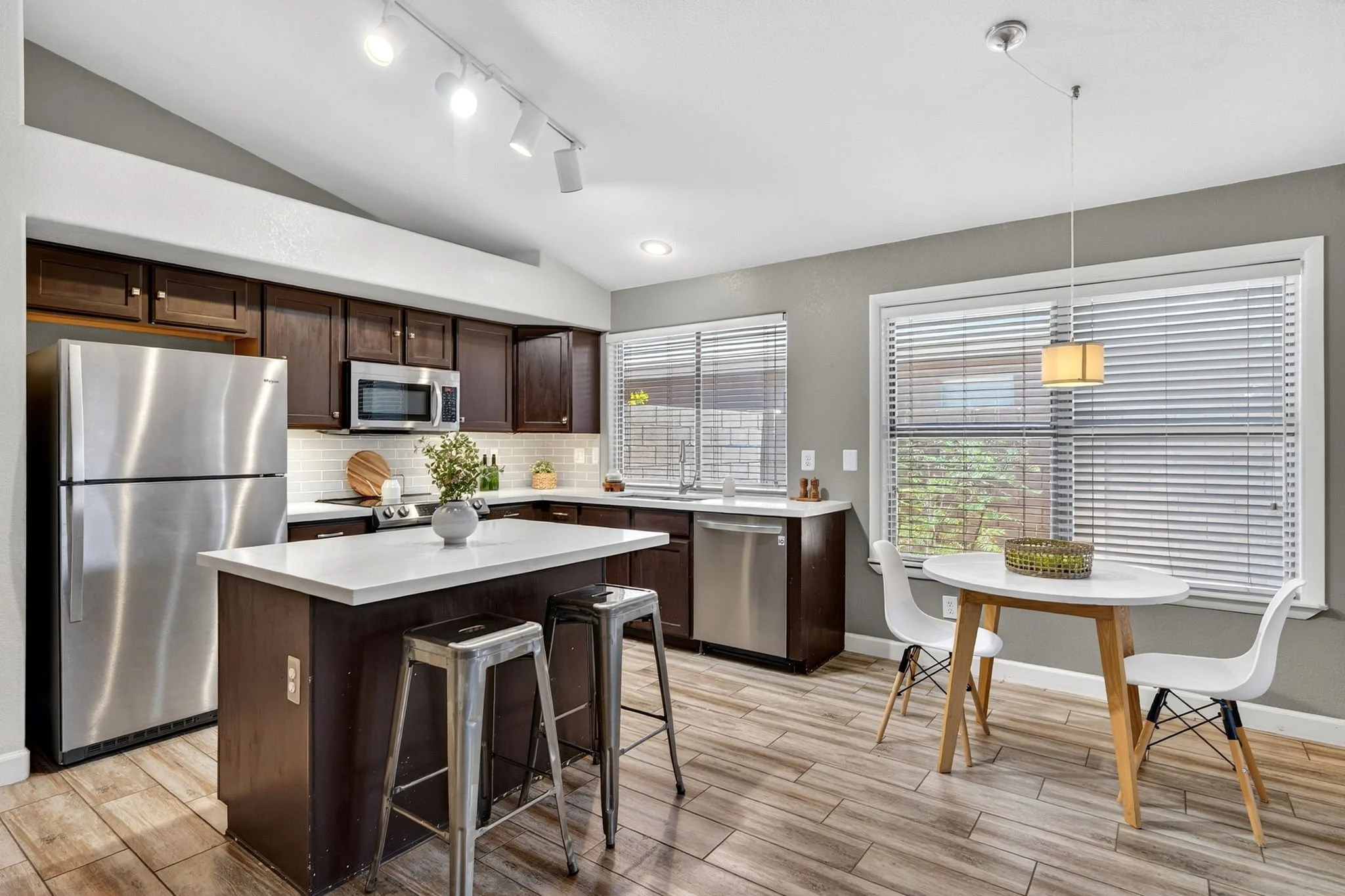 Modern kitchen with dark wooden cabinets, stainless steel refrigerator and microwave, white countertop island with stools, and a dining area with a round table and chairs near large windows with blinds.