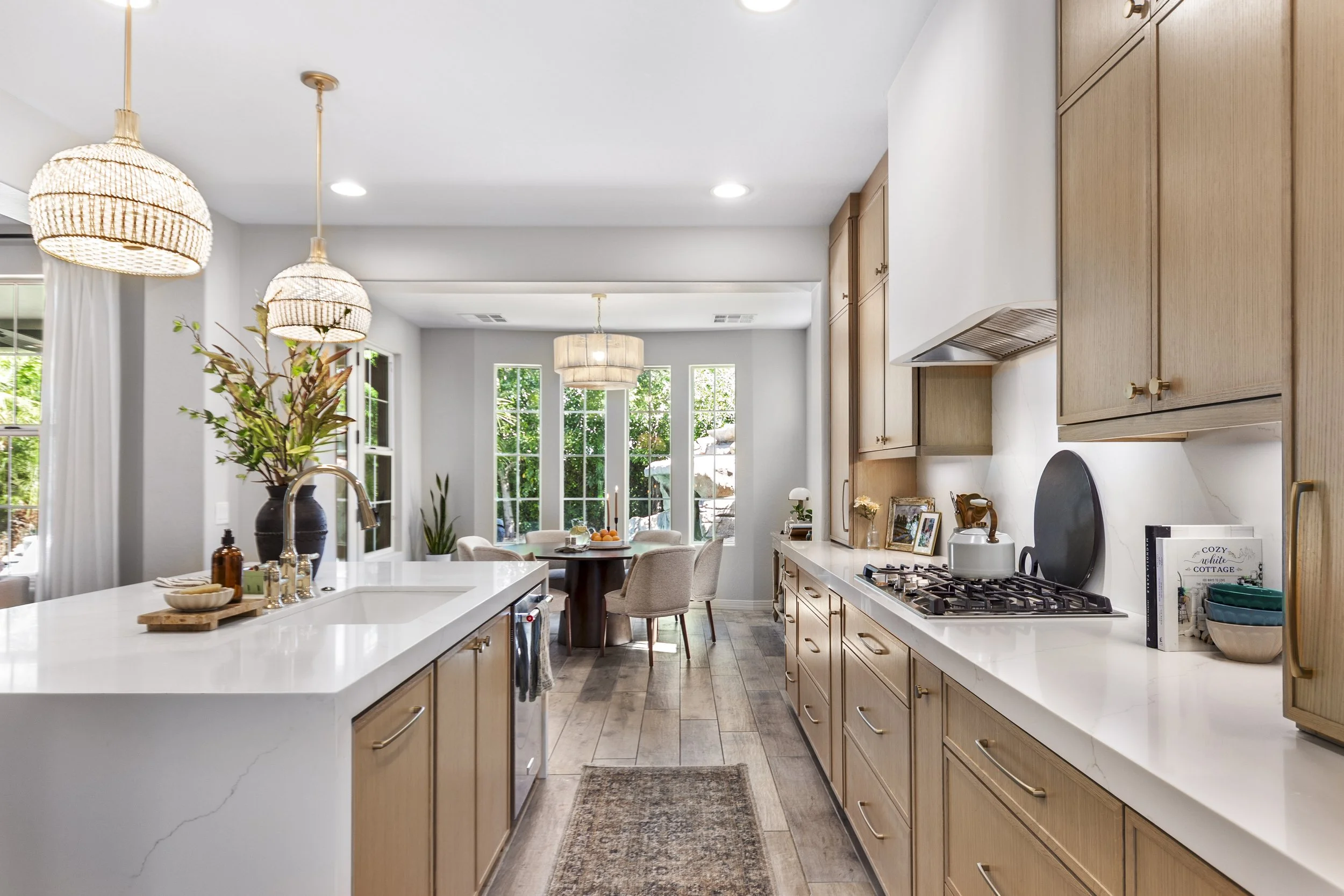Bright open kitchen with wooden cabinets, white countertops, a gas stove, and decorative elements. Visible dining area with large windows and greenery outside.