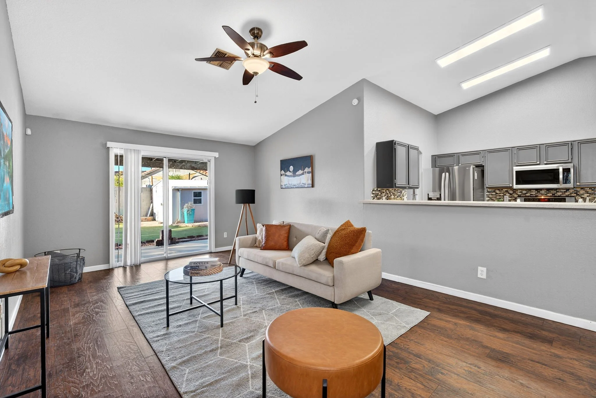 Living room with beige couch, orange pillows, black coffee table, sliding glass door, and kitchen with gray cabinets and stainless steel appliances.