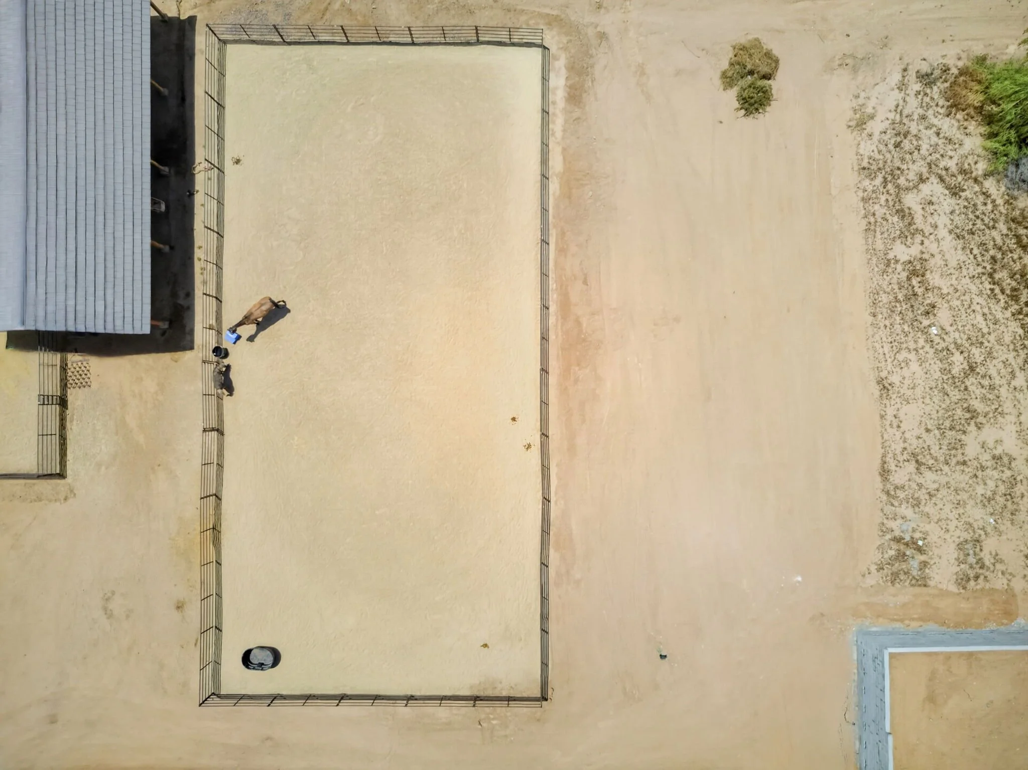 An indoor sports court with a sand or dirt surface, surrounded by a metal fence. There are a few people visible on the court, one of whom appears to be preparing to serve or hit a ball. The top of a building's roof and some sparse plants are visible 
