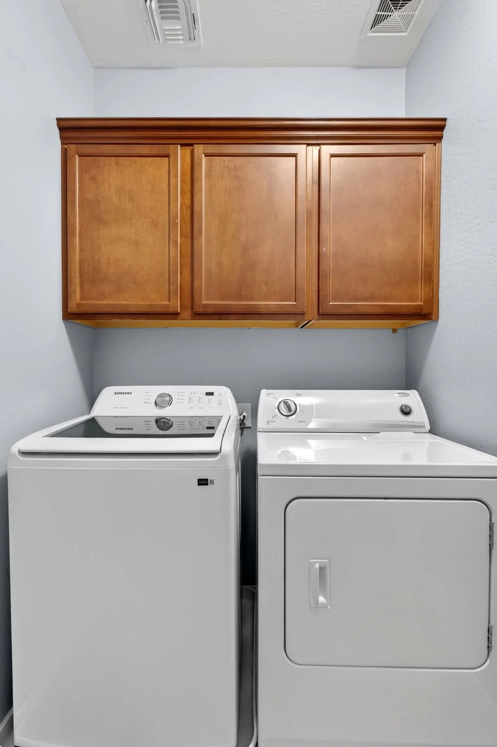 Laundry room with a Samsung washing machine on the left and a dryer on the right, above a wooden cabinet mounted on the wall.