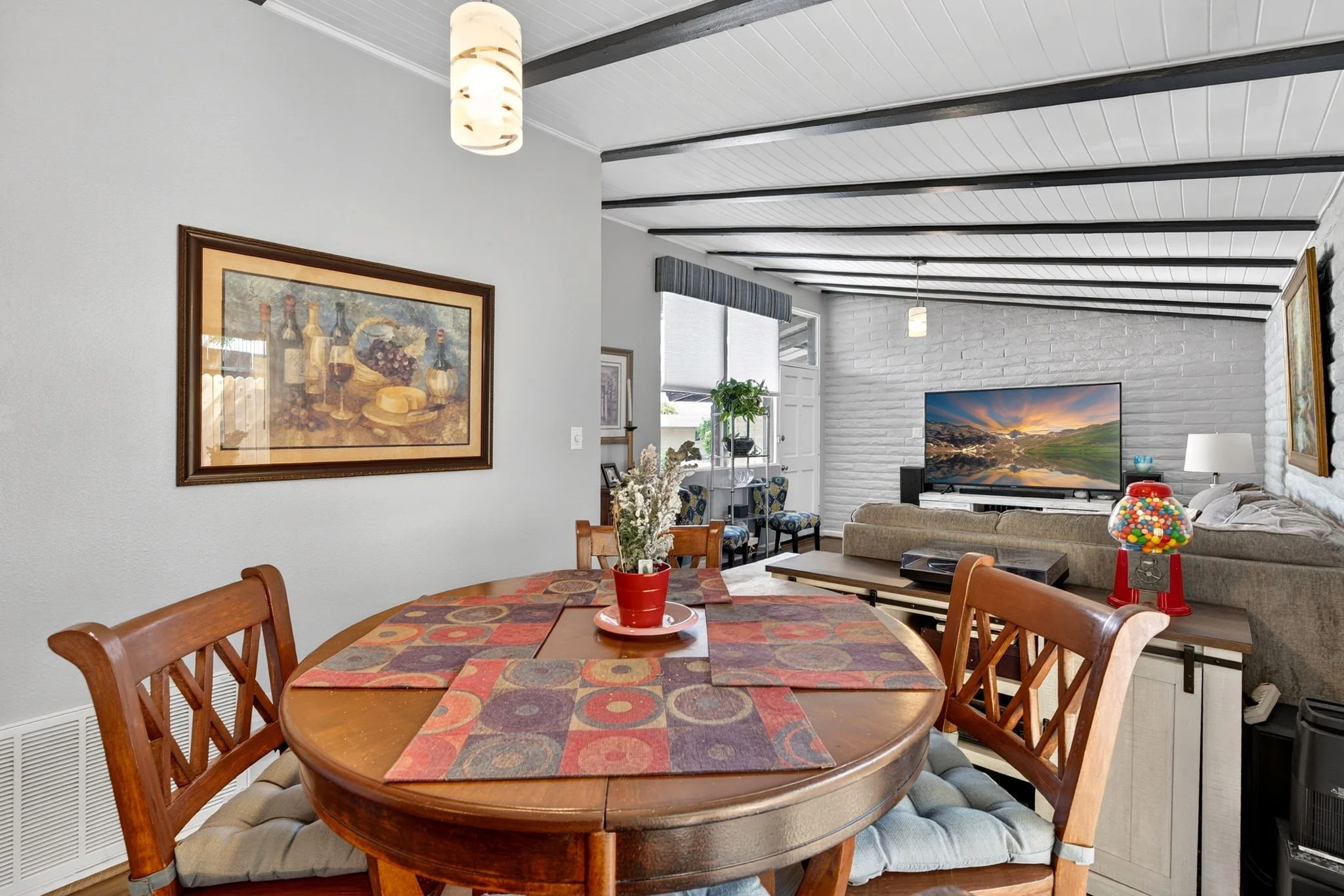 A dining area with a wooden table and six matching chairs with beige cushions. A red pot with a green plant and bouquet is on the table. The room has a white and gray color scheme with art on the walls, a white brick wall, a flat-screen TV, and a gum