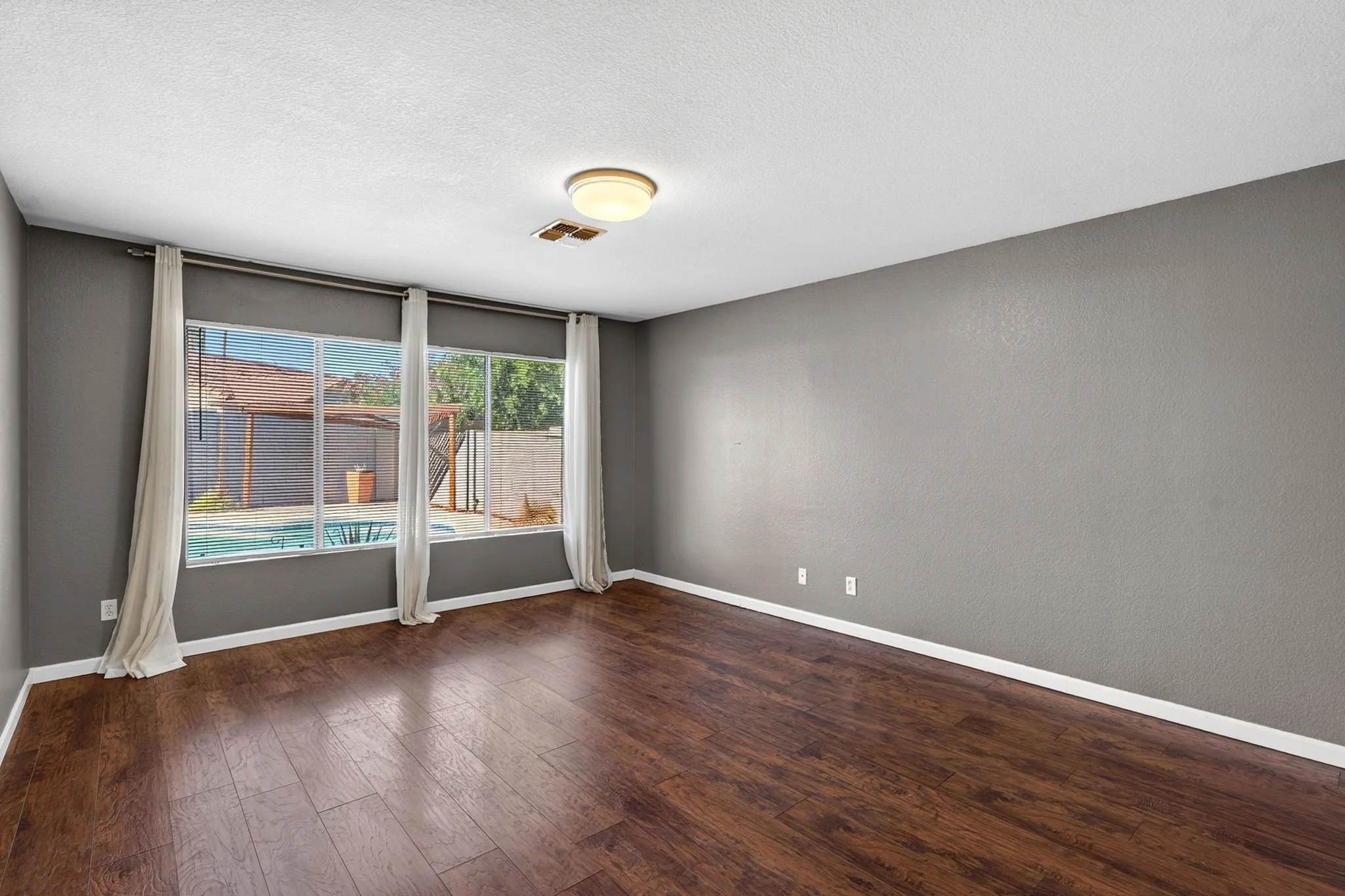 Empty living room with brown hardwood floors, gray walls, large window with white curtains, and a ceiling light fixture.