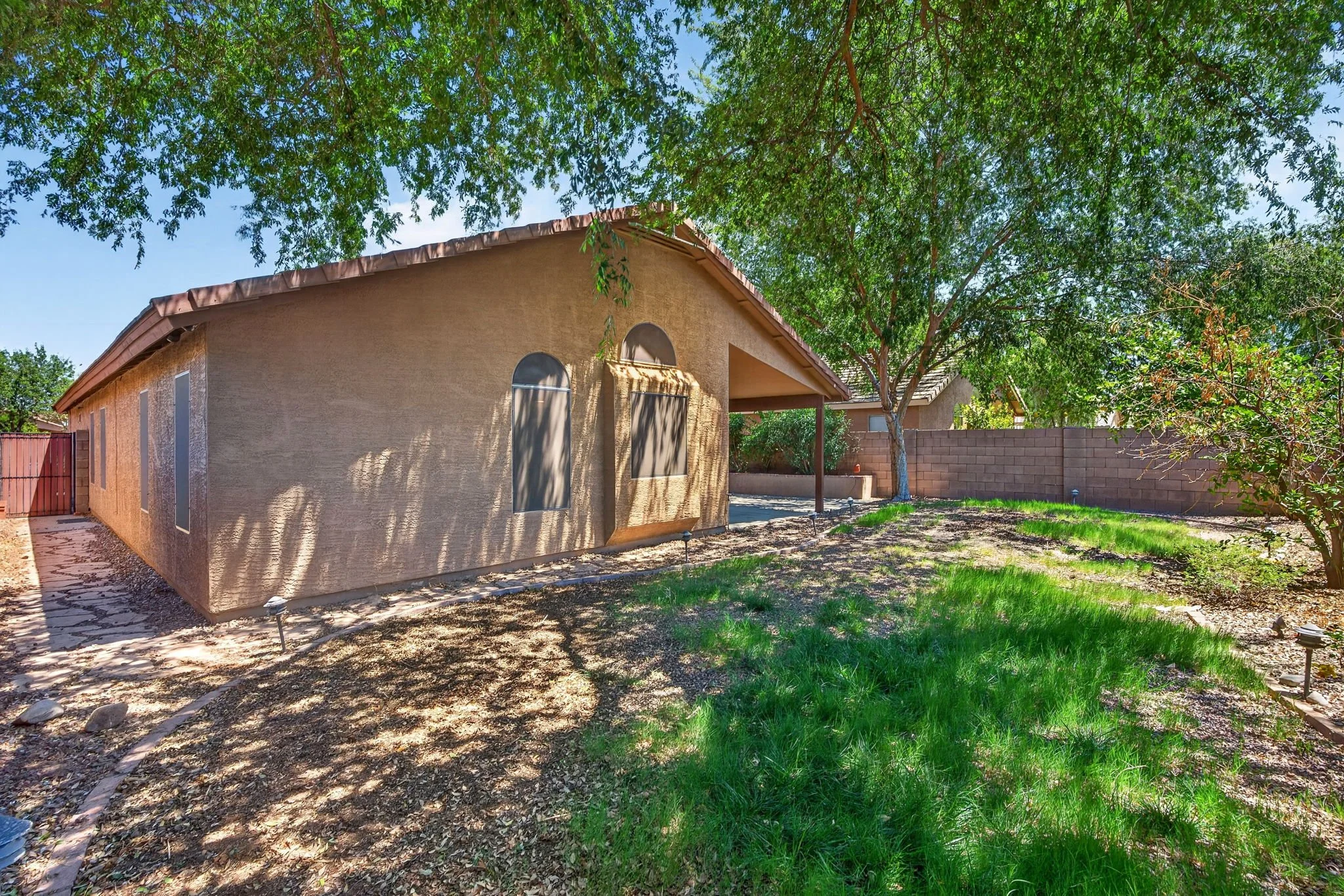 Exterior view of a stucco house with a brown tile roof, surrounded by a backyard with green grass, trees, and a brick wall fence.